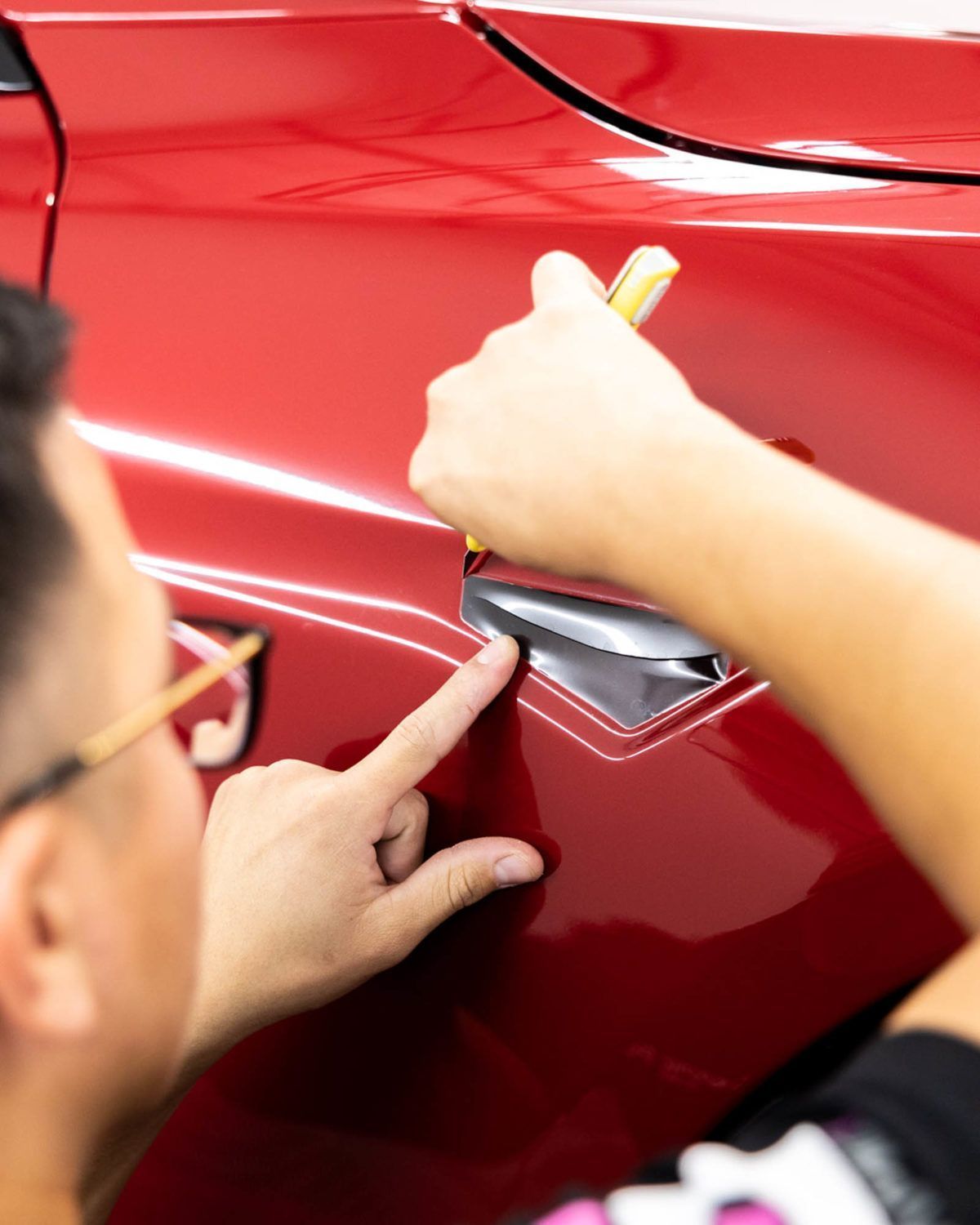 A person carefully applies a gray automotive vinyl wrap to the door handle area of a red car.