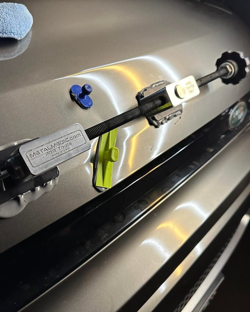 A paintless dent repair tool pulling a dent on a car hood, with a blue glue tab and green plastic wedge nearby.