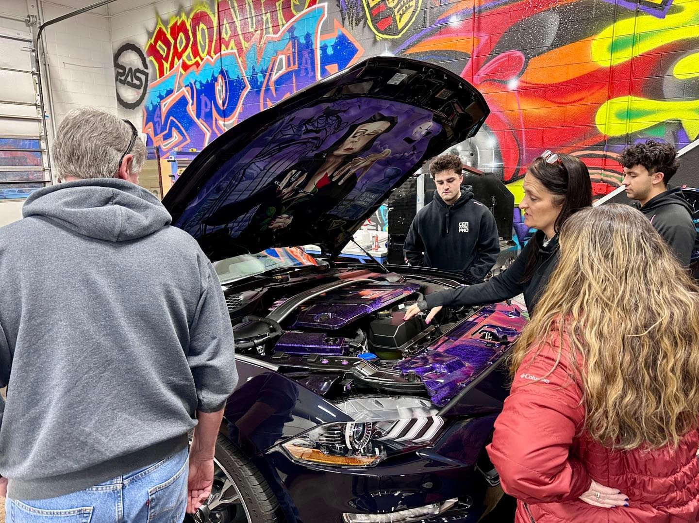 A group of people observe a custom, purple-painted car with an open hood in a garage decorated with colorful graffiti.