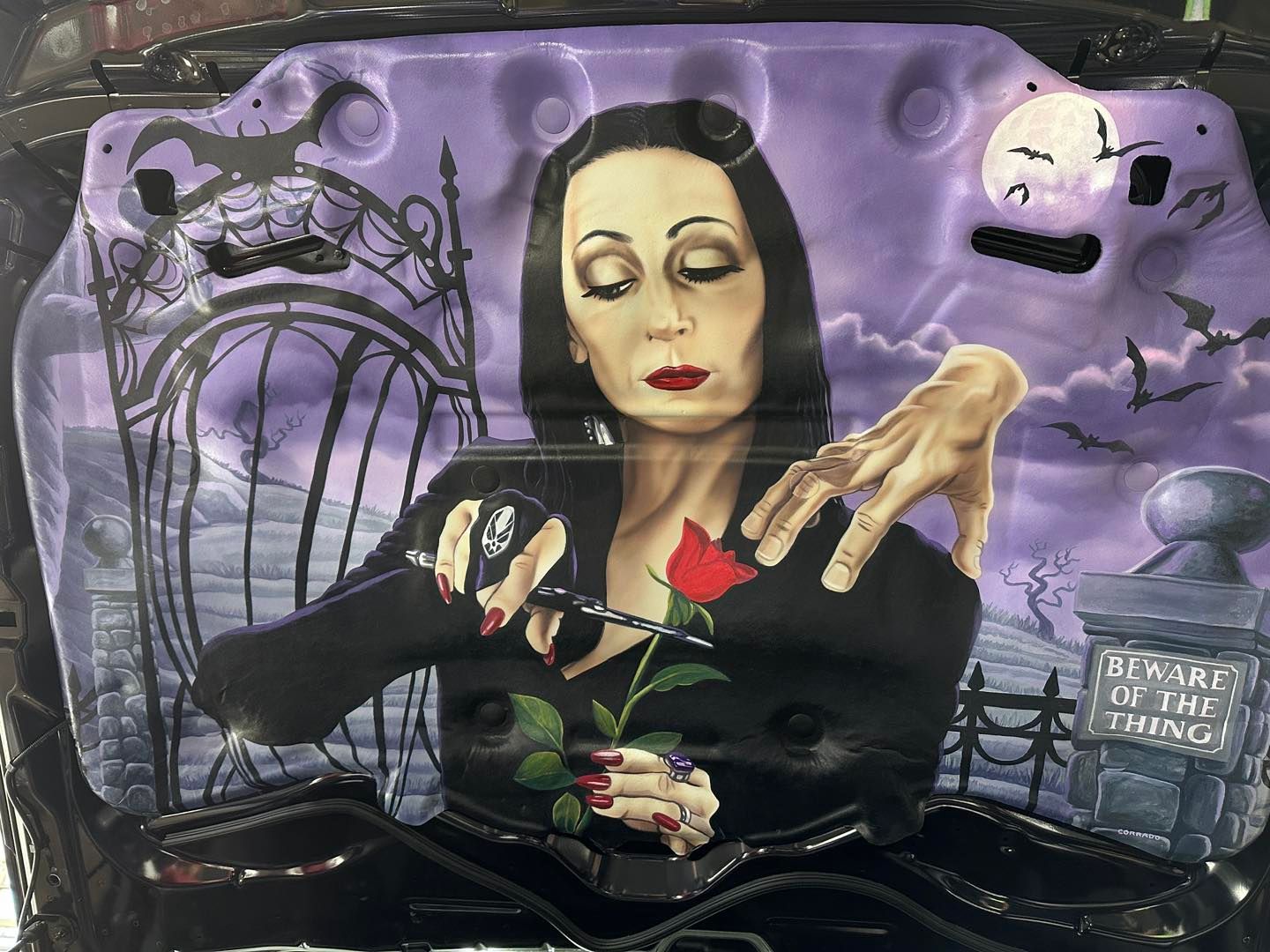 Airbrushed art of Morticia Addams snipping a rose, set against a spooky purple graveyard with bats and a full moon.