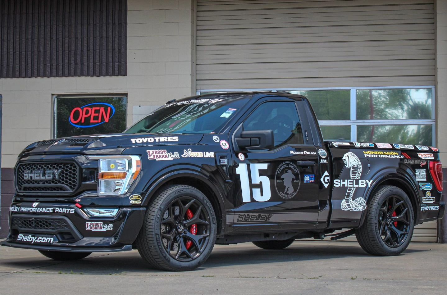 Black Shelby F-150 truck with racing decals, number 15, parked in front of a building with an open sign.