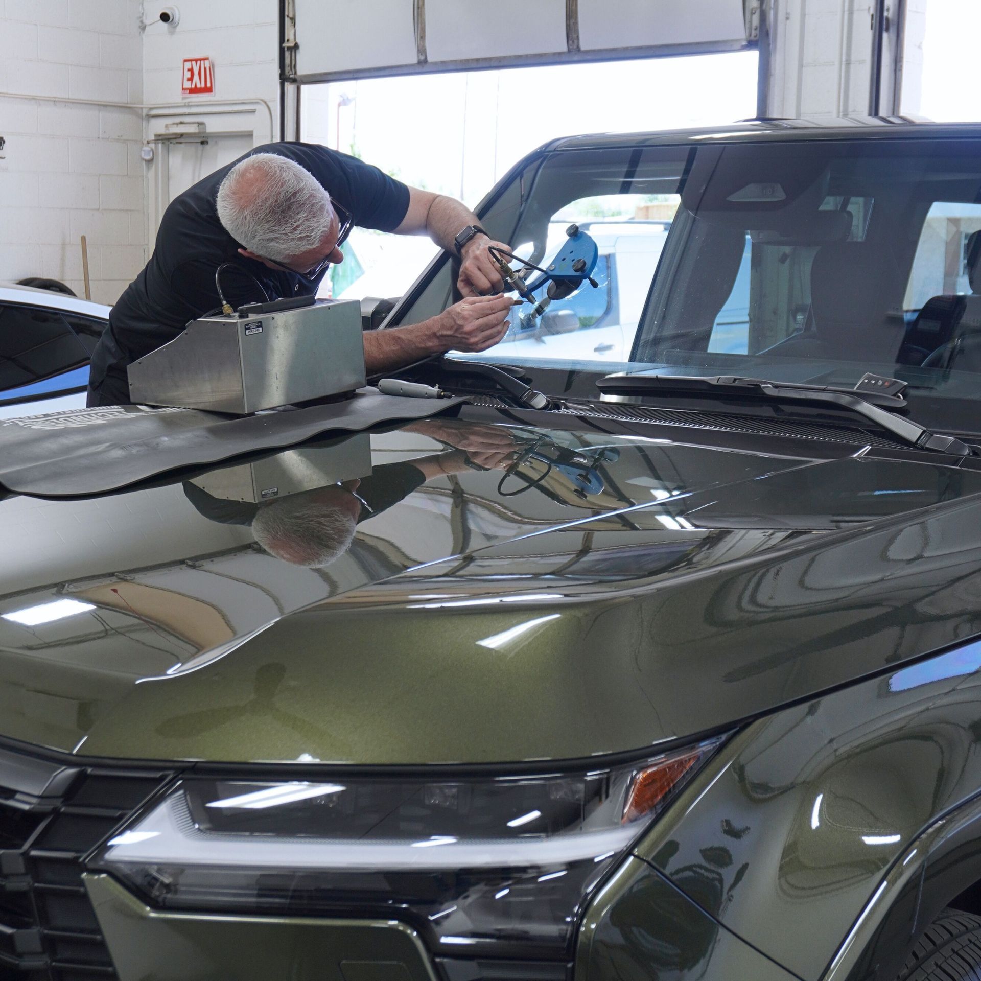 A technician repairs a windshield chip on an olive-green SUV in a garage.