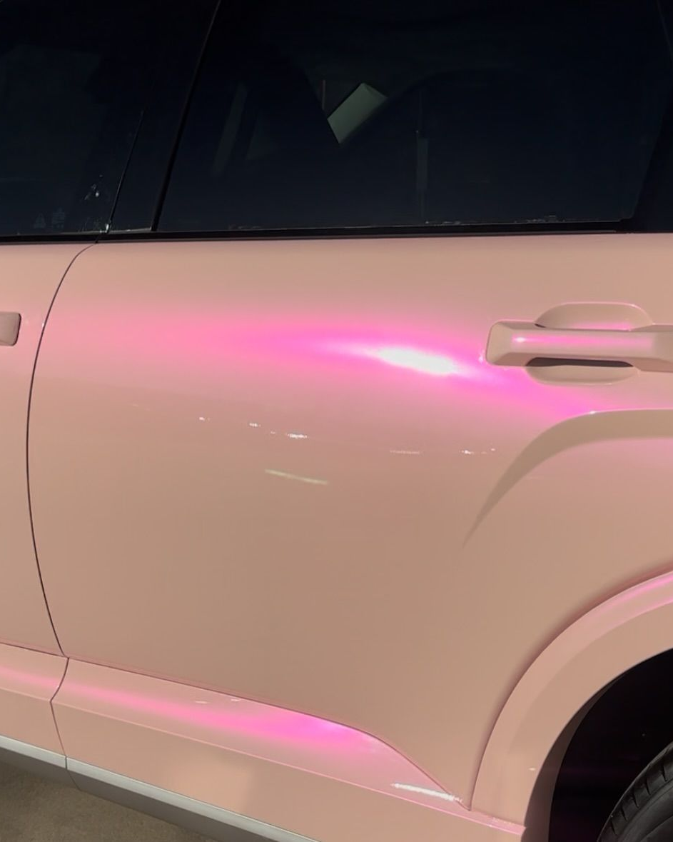 A person in work clothes and gloves uses a tool to apply a bronze-colored vinyl wrap to a car door.