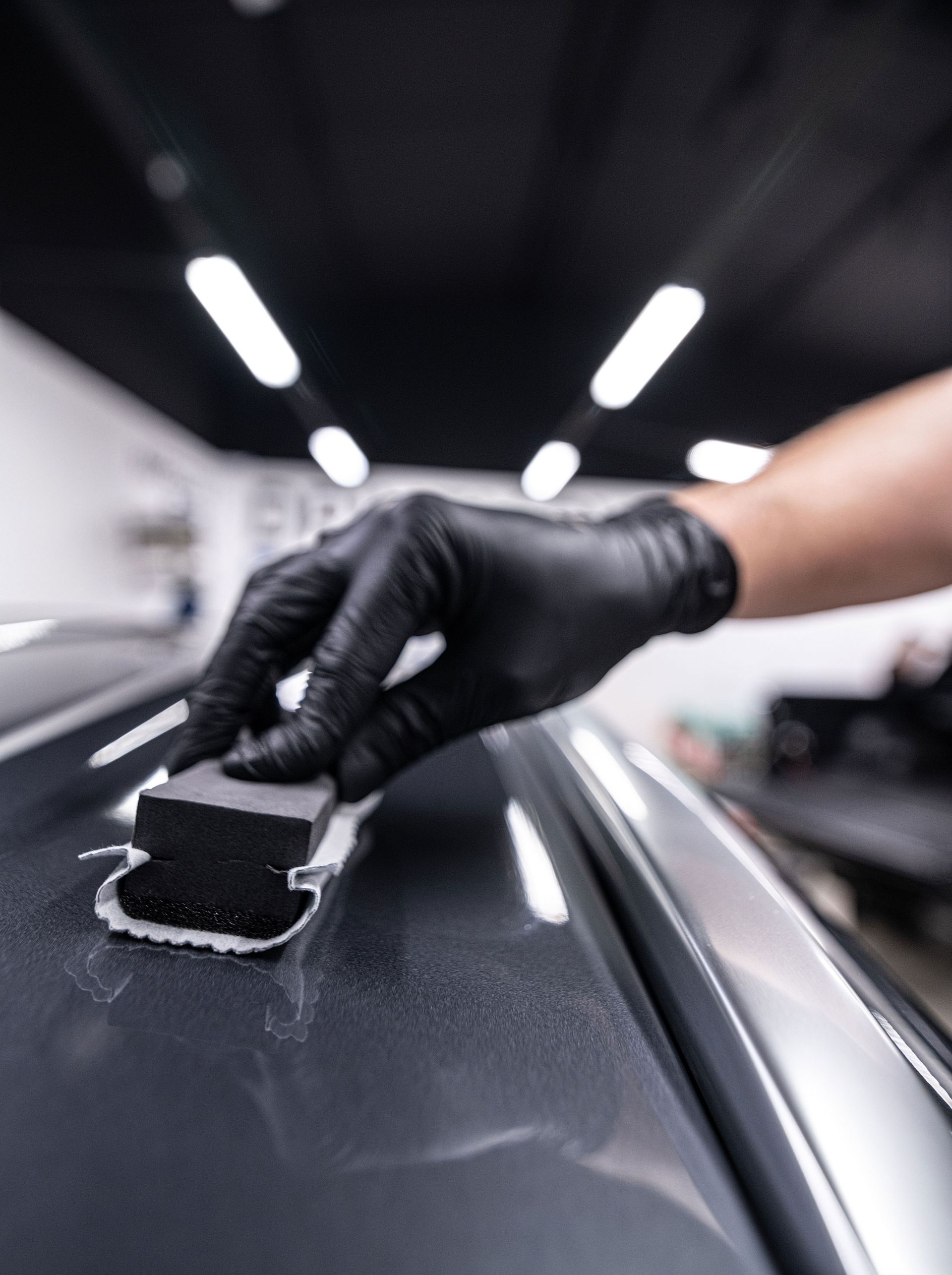 A person wearing black nitrile gloves applies a ceramic coating to a gray car panel using a foam applicator block.