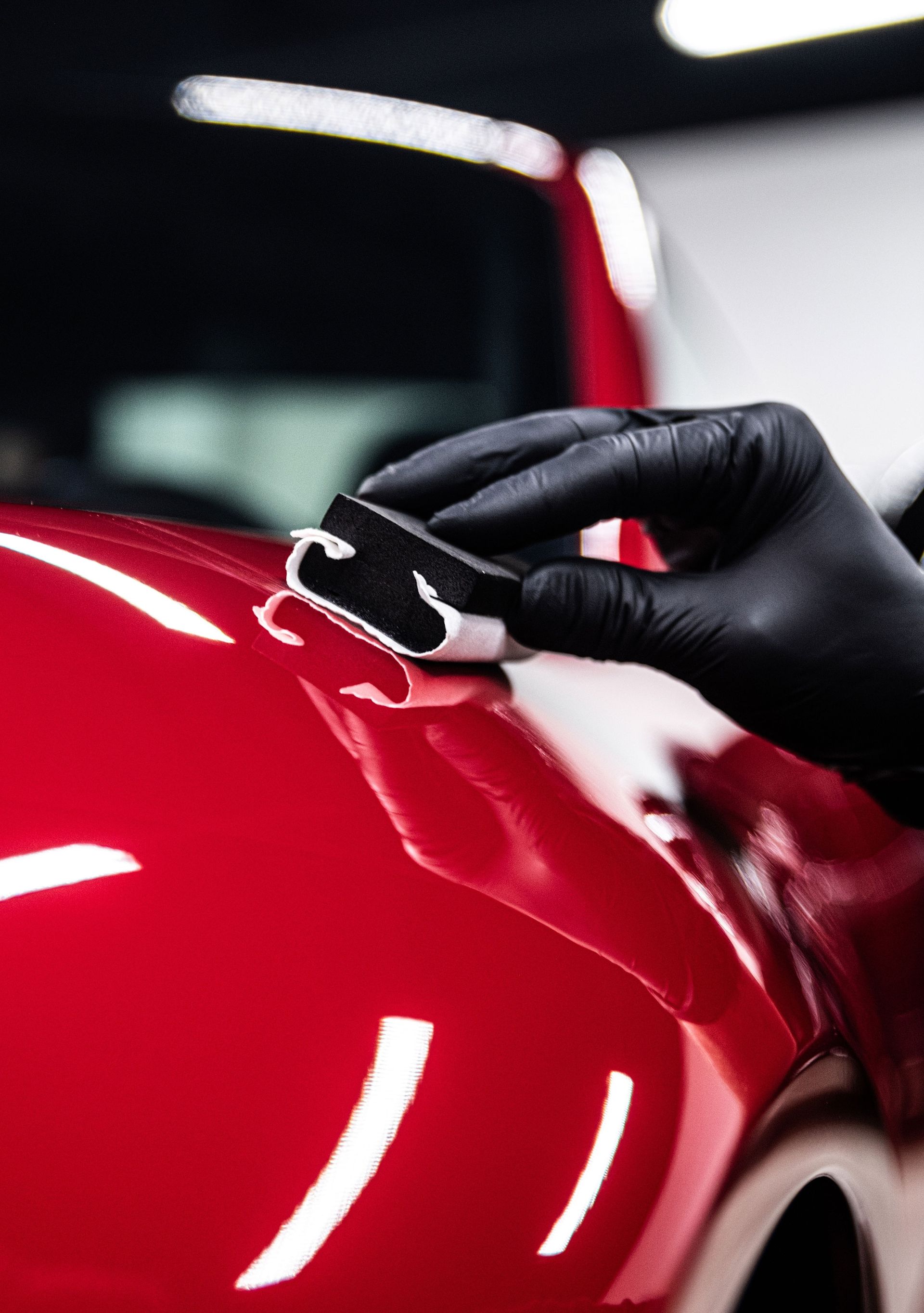 A gloved hand uses a foam block applicator to apply a ceramic coating to a glossy red car panel.