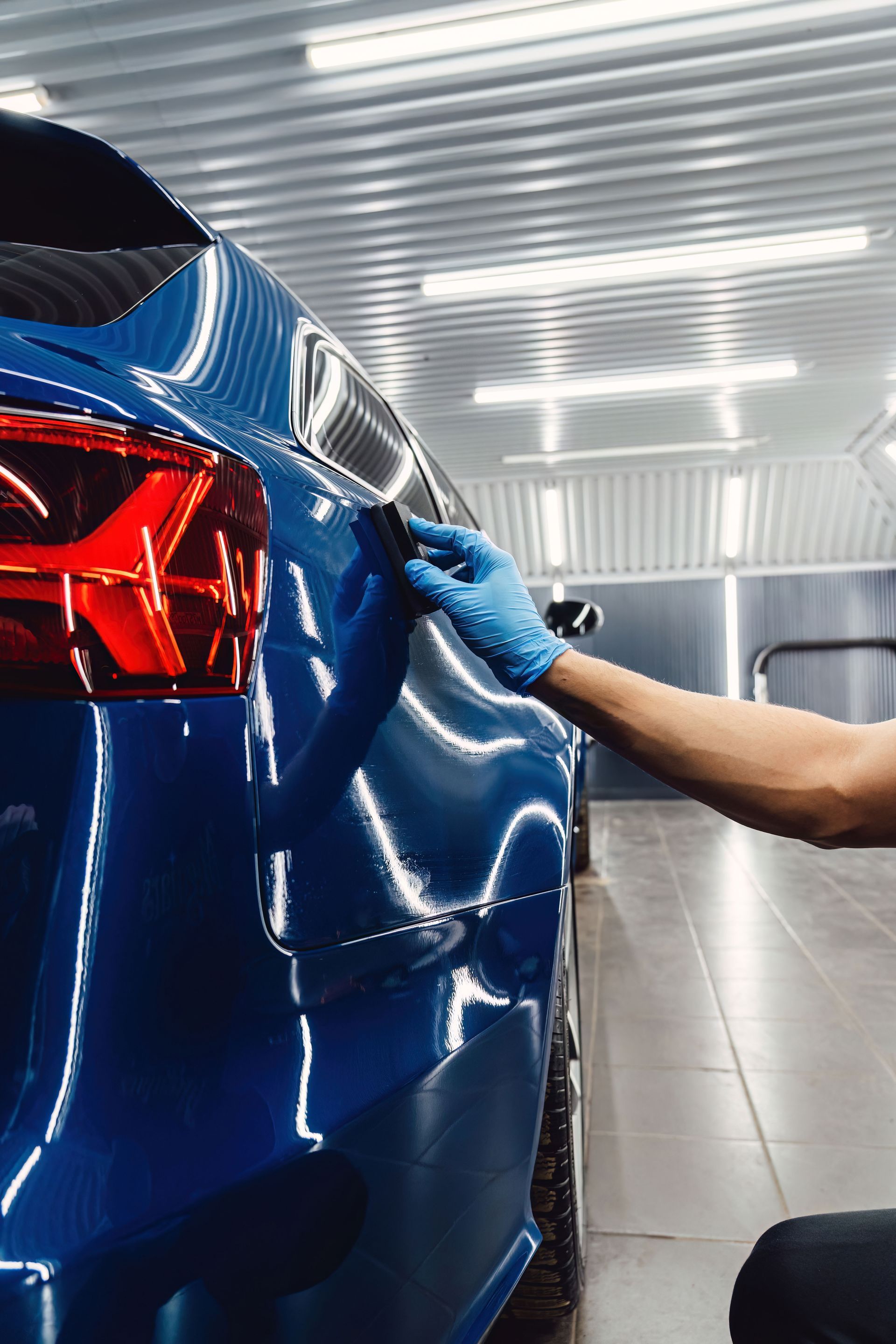 A gloved hand uses a detailing pad to apply a protective coating to the blue rear quarter panel of a car in a shop.