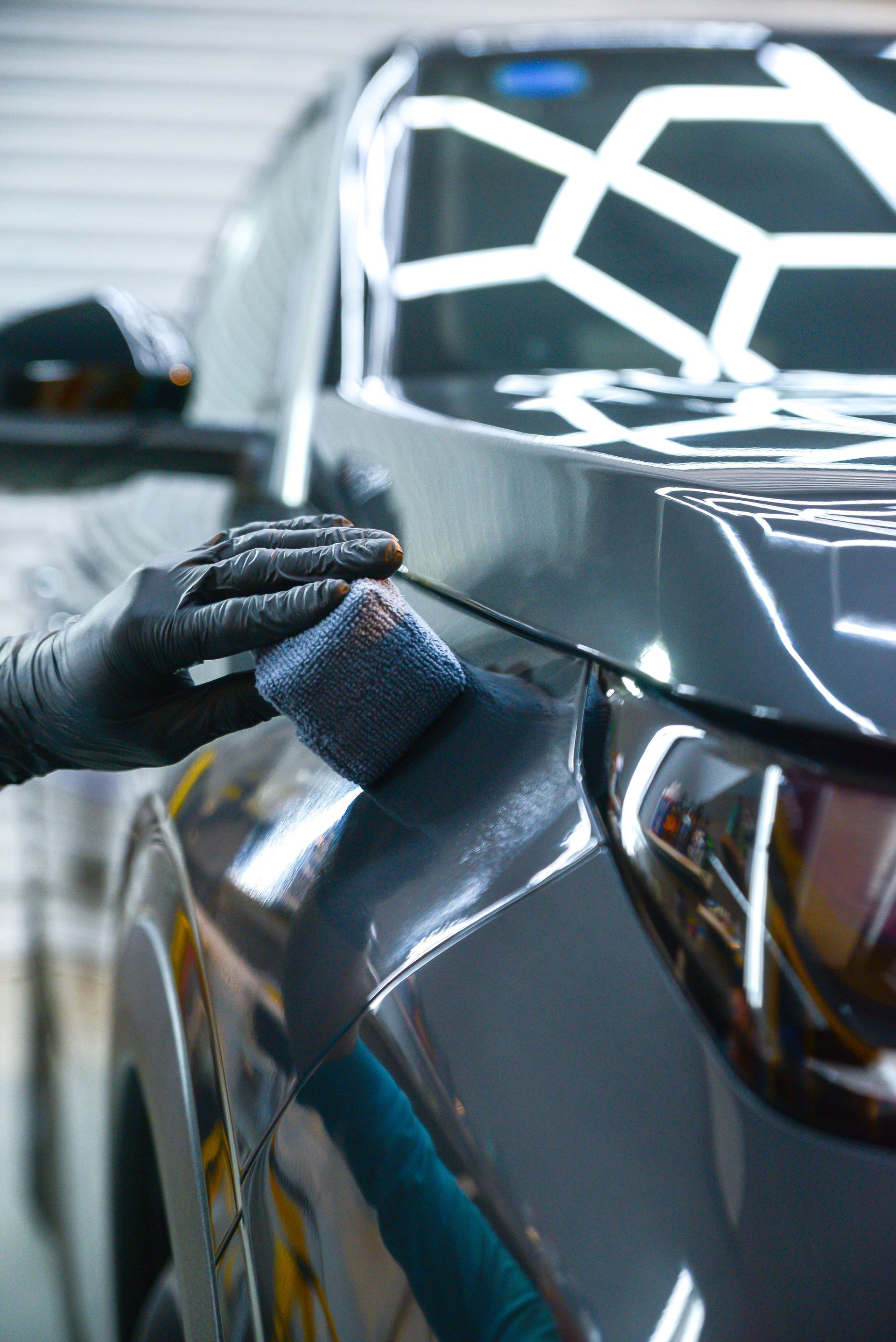 A hand in a black glove applies a ceramic coating to a dark gray car using a small applicator pad.