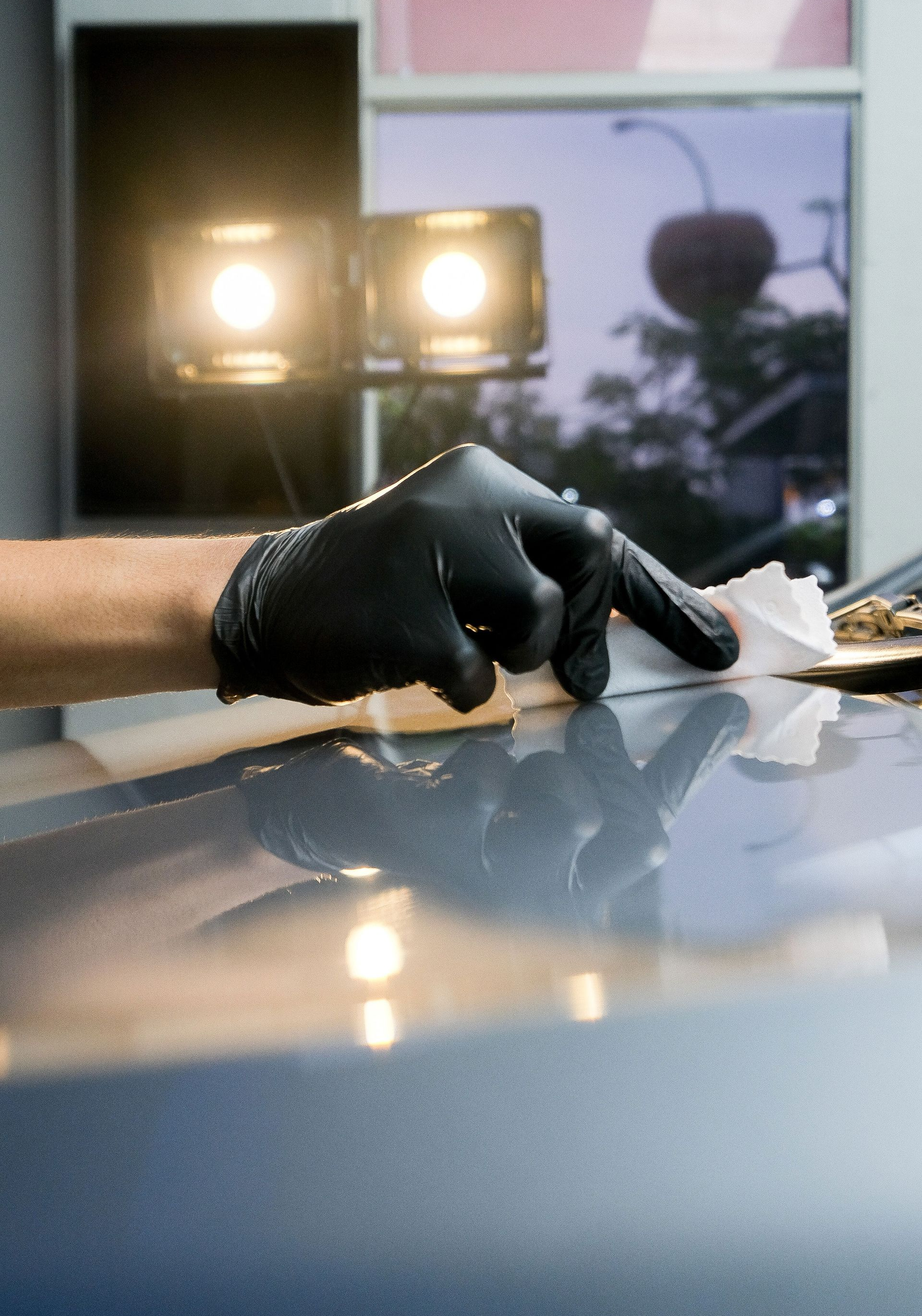 A hand in a black glove uses a white cloth to wipe the surface of a polished, blue car hood under bright studio lights.