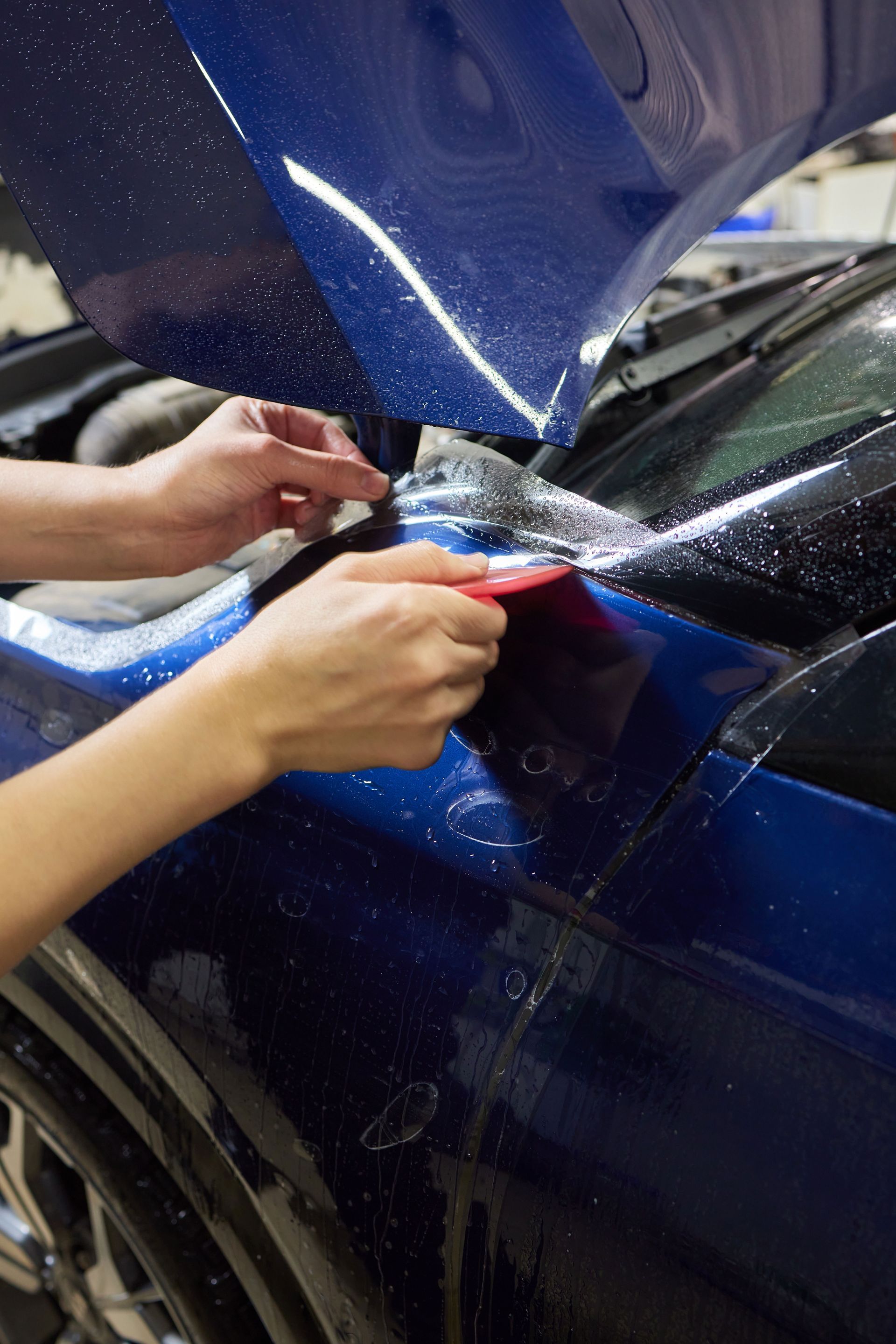 Hands use a red squeegee to apply a clear protective film onto the blue fender of a vehicle.