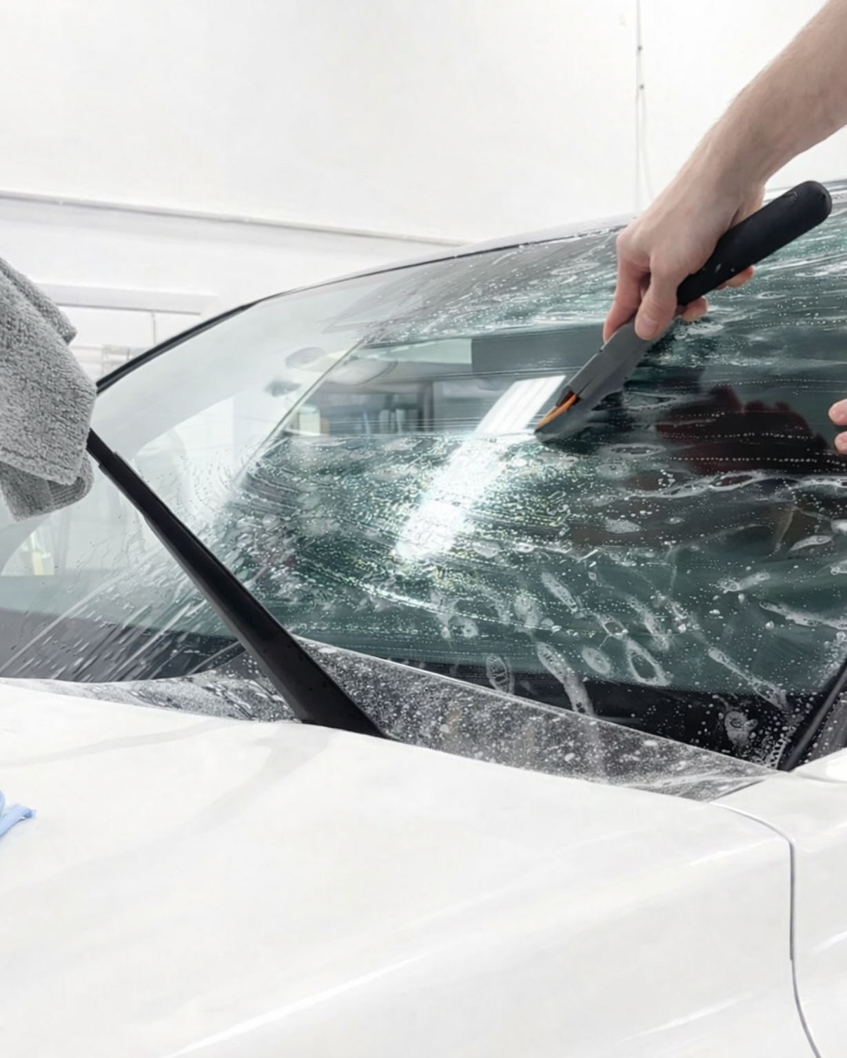 A person uses a squeegee to smooth window tint film onto a car windshield in an indoor garage.