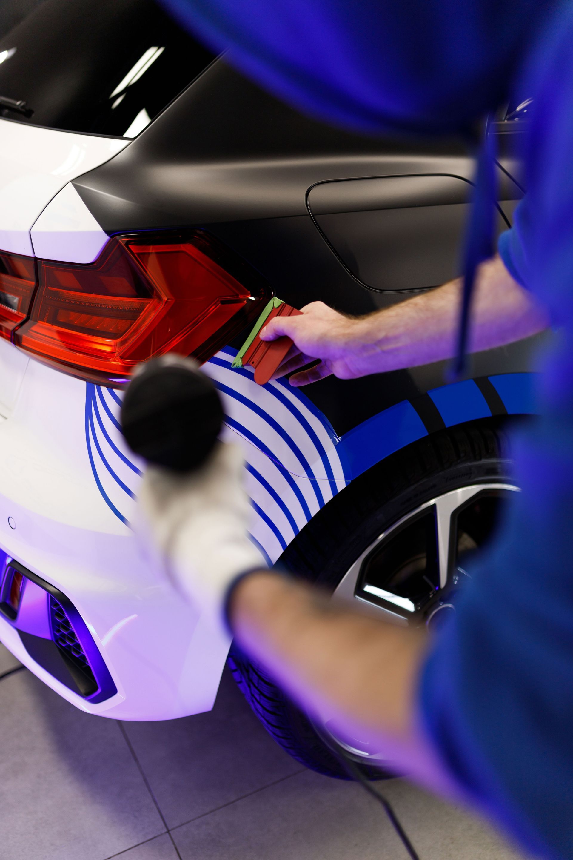 A person wearing a glove and using a heat gun and squeegee to apply a vinyl wrap to a car's rear panel.