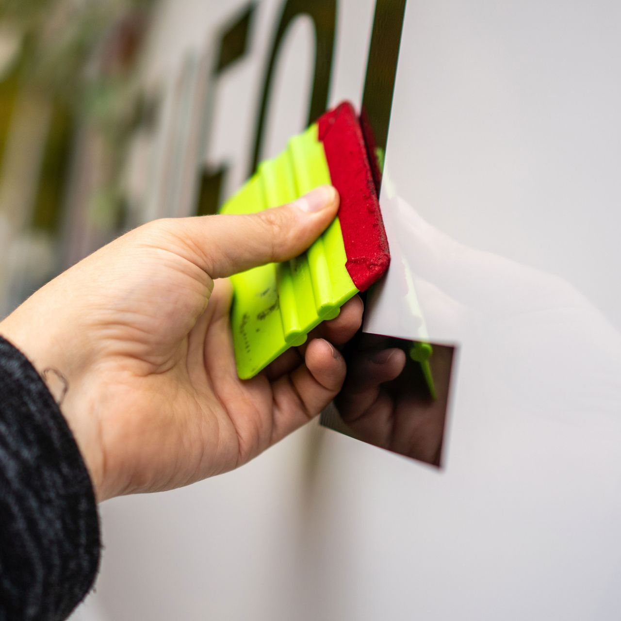 A hand uses a green and red squeegee to smooth a vinyl sticker onto a white surface.