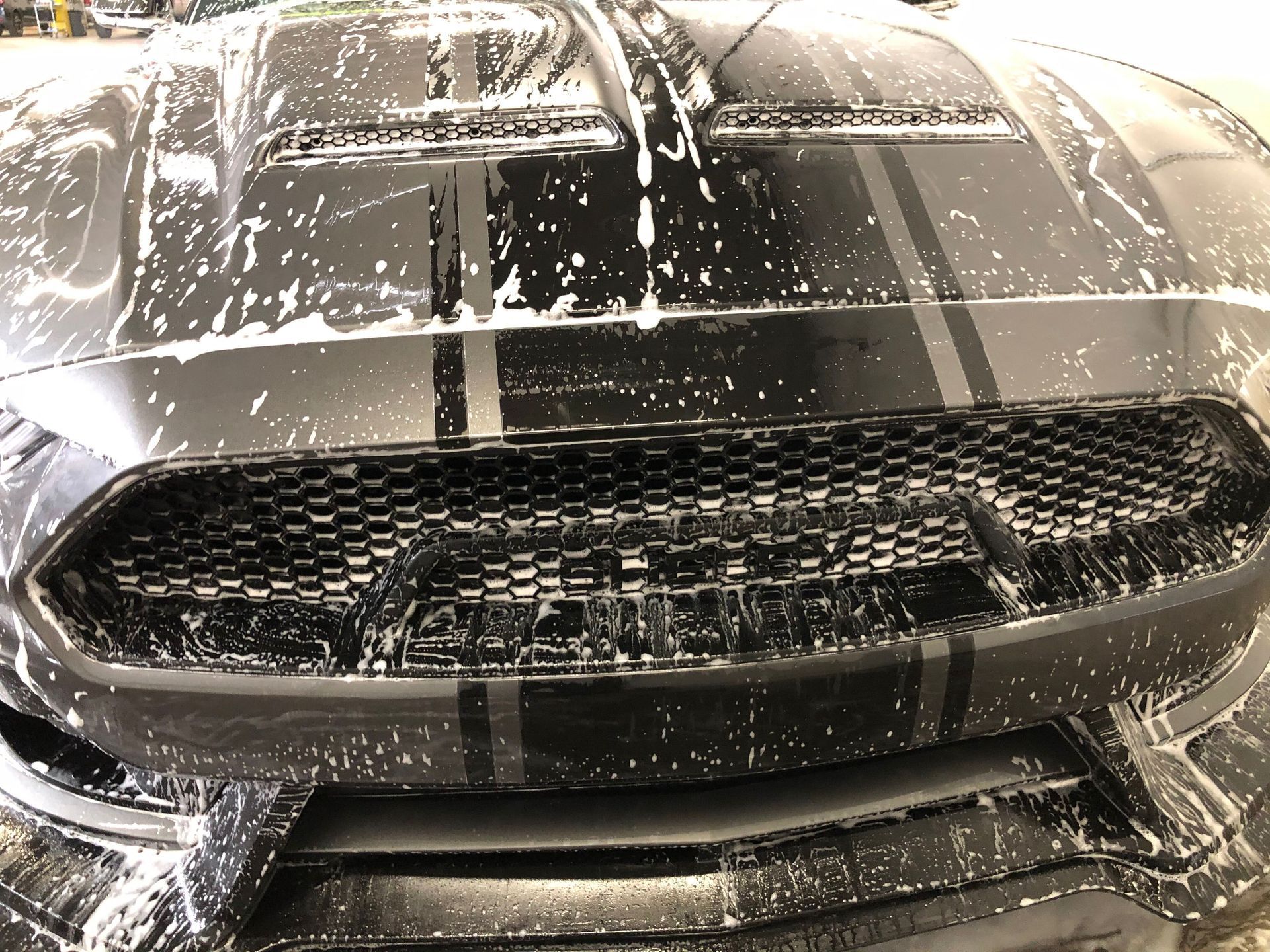A close-up of a black car with racing stripes covered in white soap suds during a car wash.