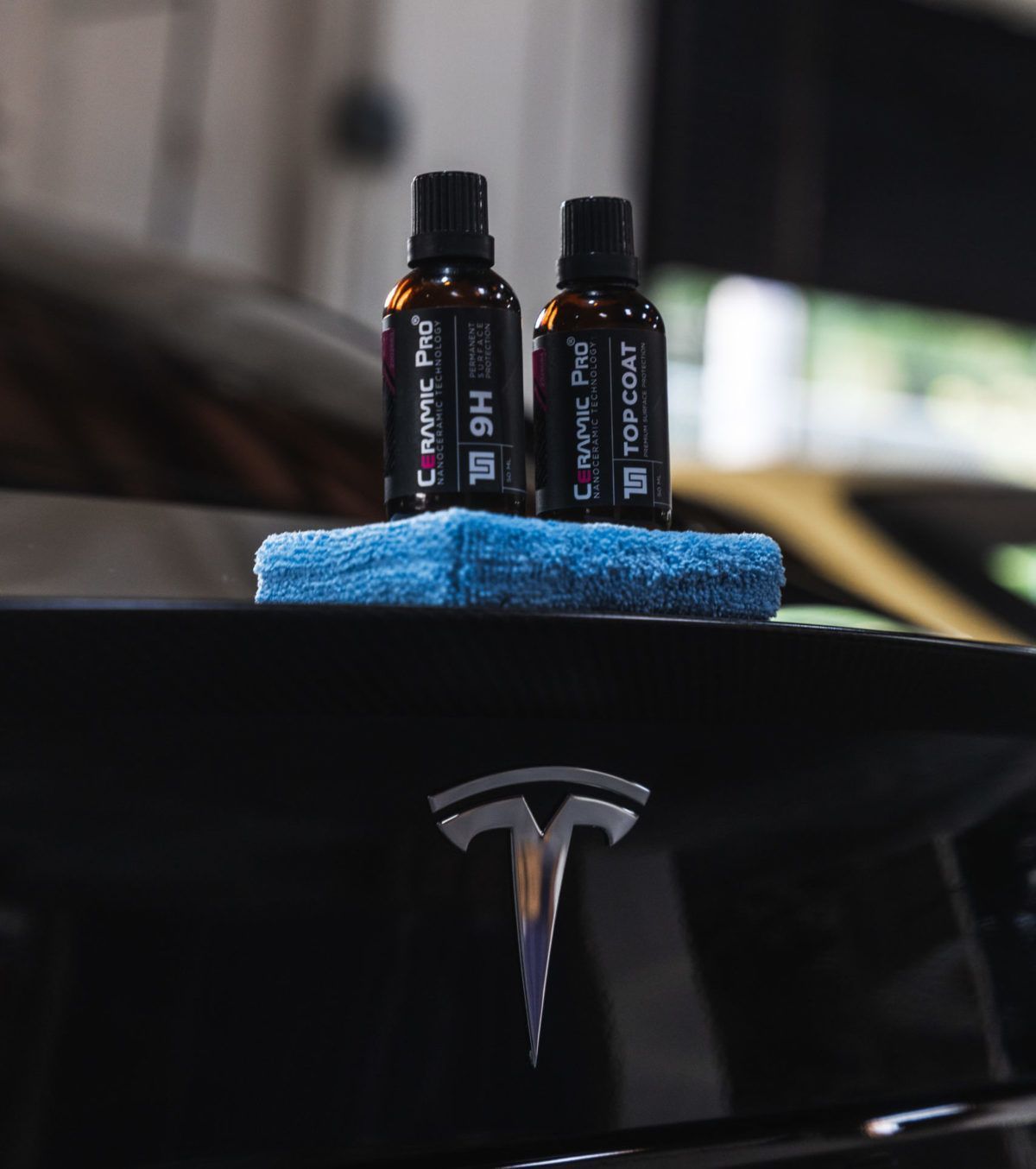 Two bottles of Ceramic Pro coating and a blue applicator pad resting on the shiny black hood of a Tesla.