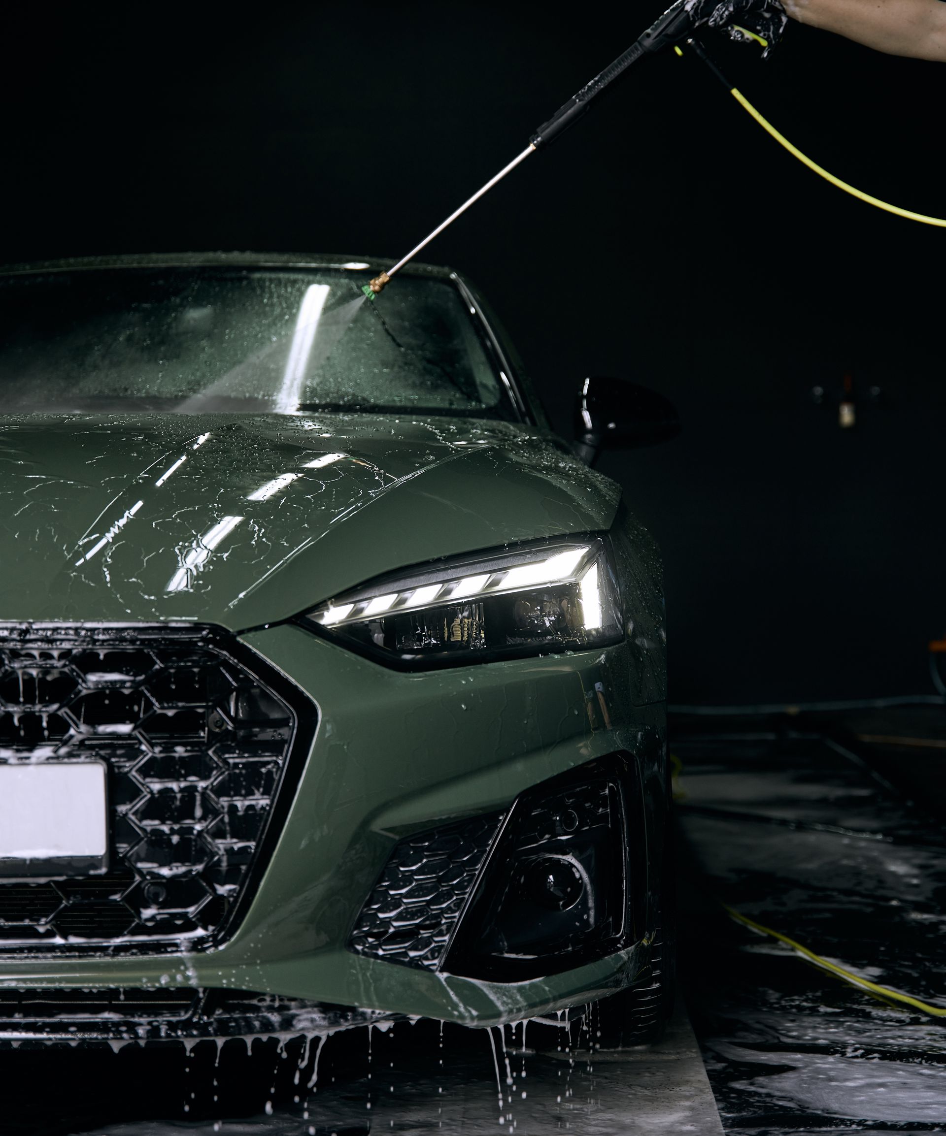 A green car being spray washed with water and soap against a dark, indoor background.