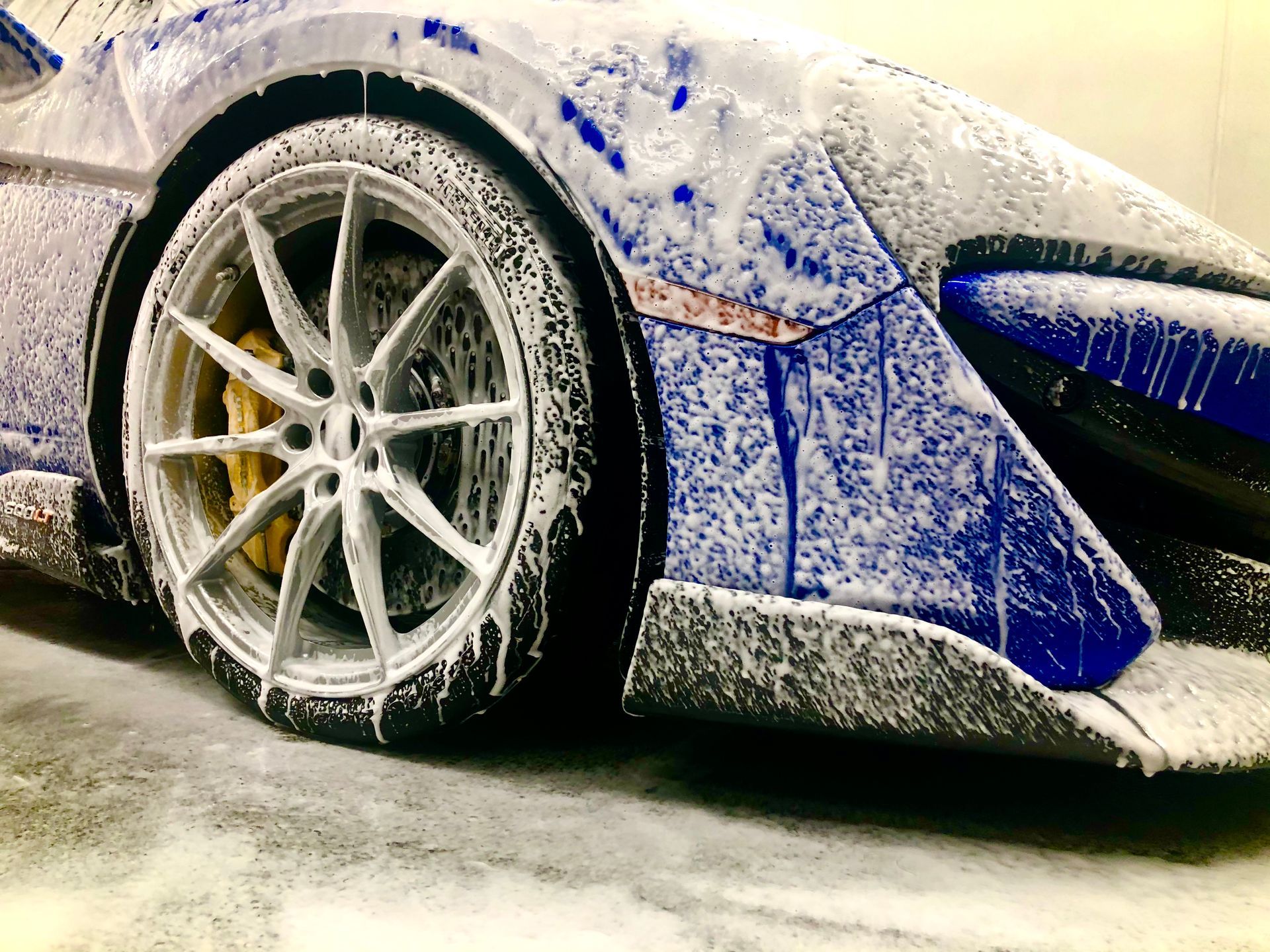 A close-up view of a blue sports car covered in thick white soap suds during a car wash.