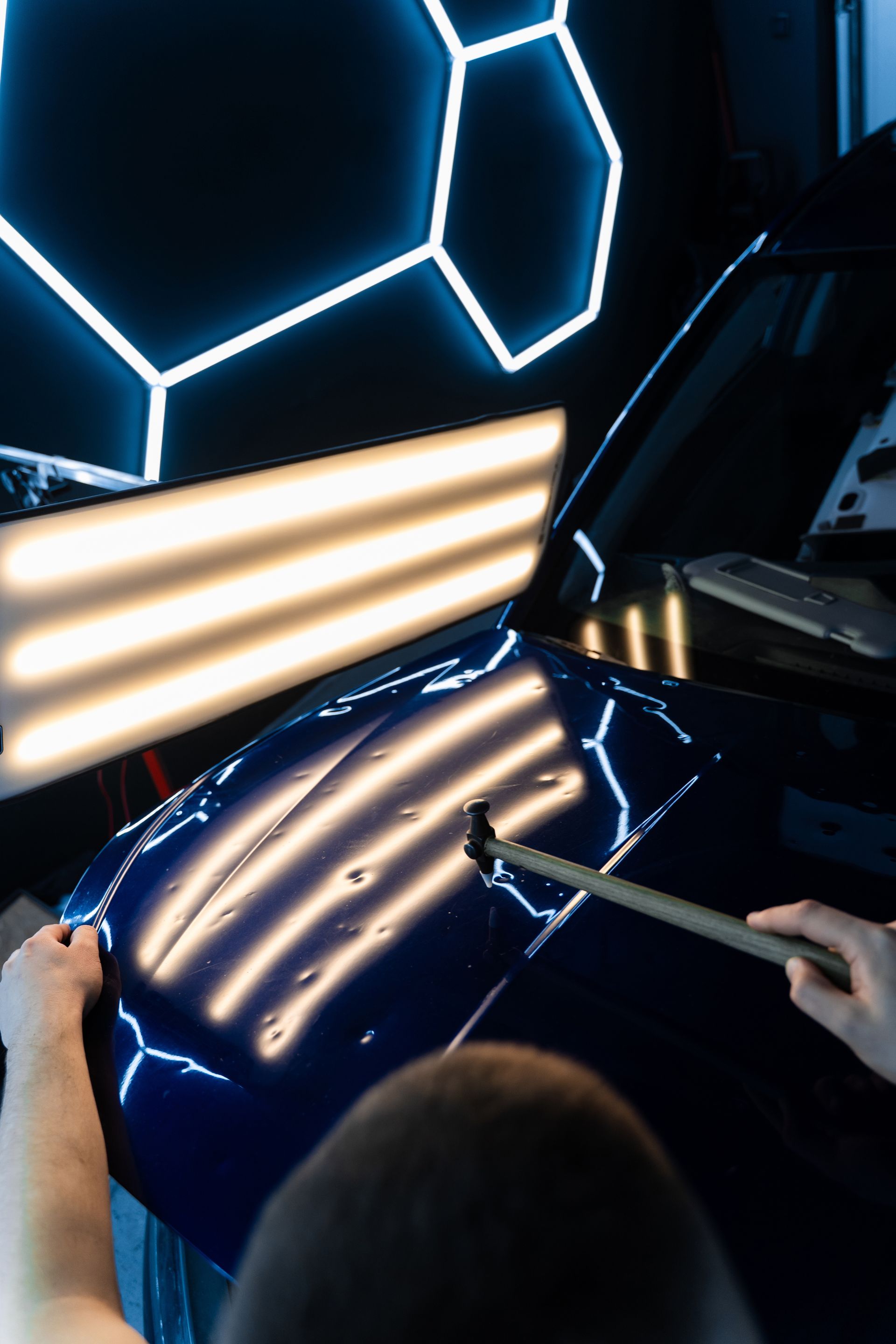 A person uses a specialized tool to perform paintless dent repair on a dark blue car hood under bright, striped light panels.