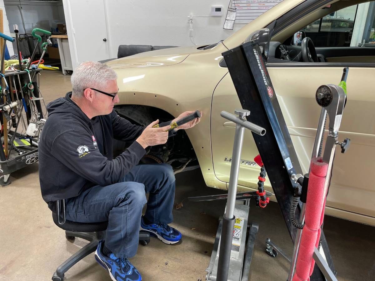 A technician works on the front fender of a beige car in a workshop, using a specialized tool for dent repair.