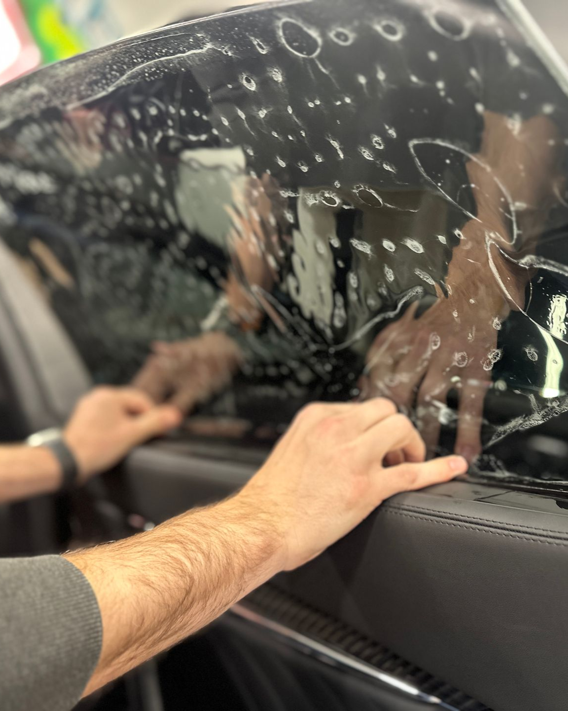 Hands peeling clear protective film from a car window inside a vehicle.
