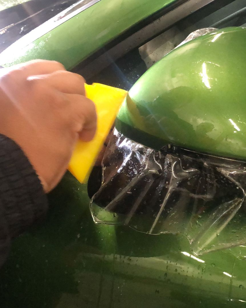 Man installing paint protection film on a red car in a garage, using a headlamp.