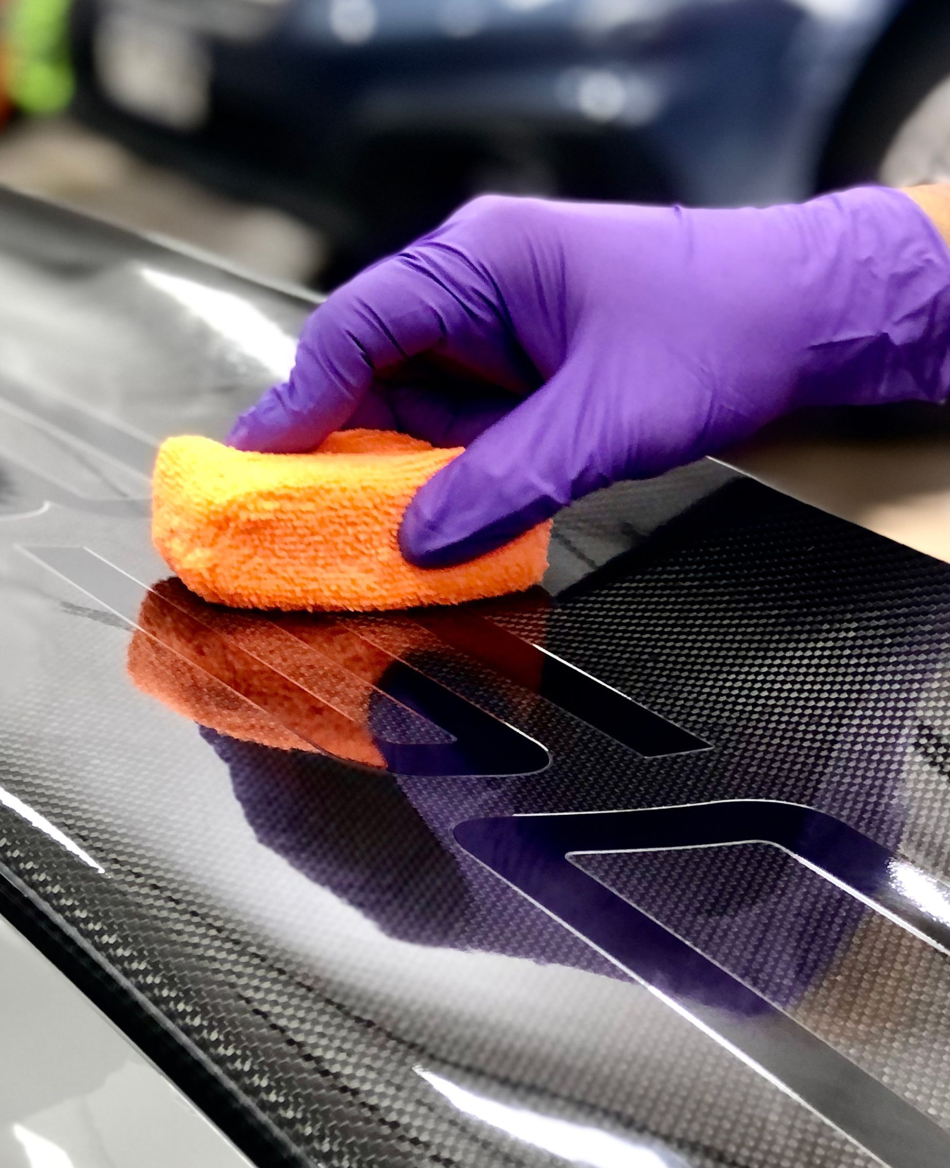 A hand in a purple nitrile glove uses an orange sponge to apply product to a dark, carbon fiber car surface.