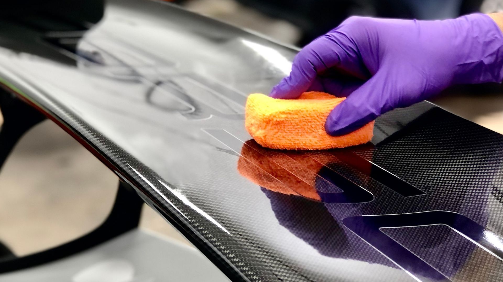A hand in a purple nitrile glove uses an orange sponge to apply a coating to a black carbon fiber vehicle spoiler.