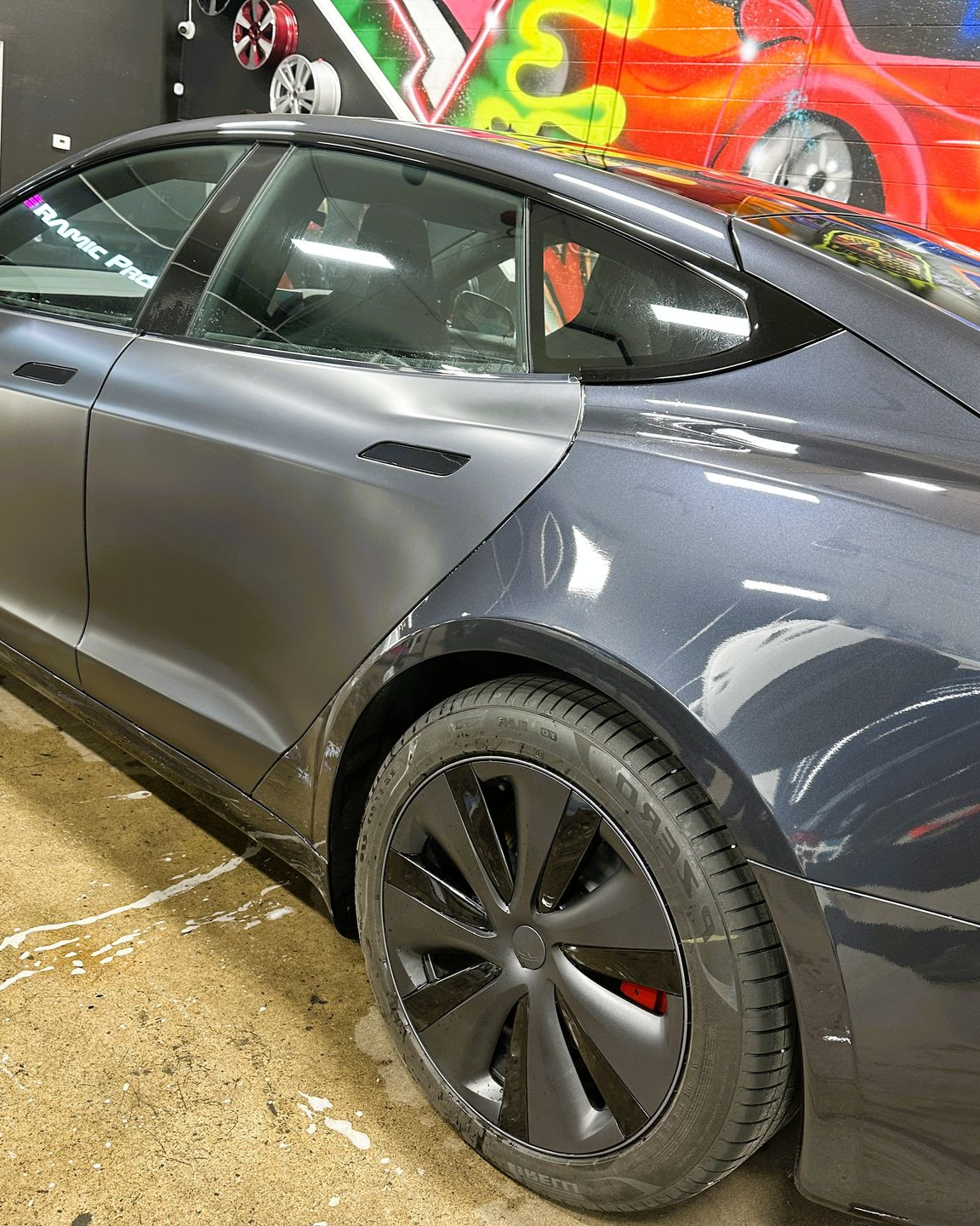 Gray Tesla sedan parked indoors with black wheels in front of a colorful graffiti wall
