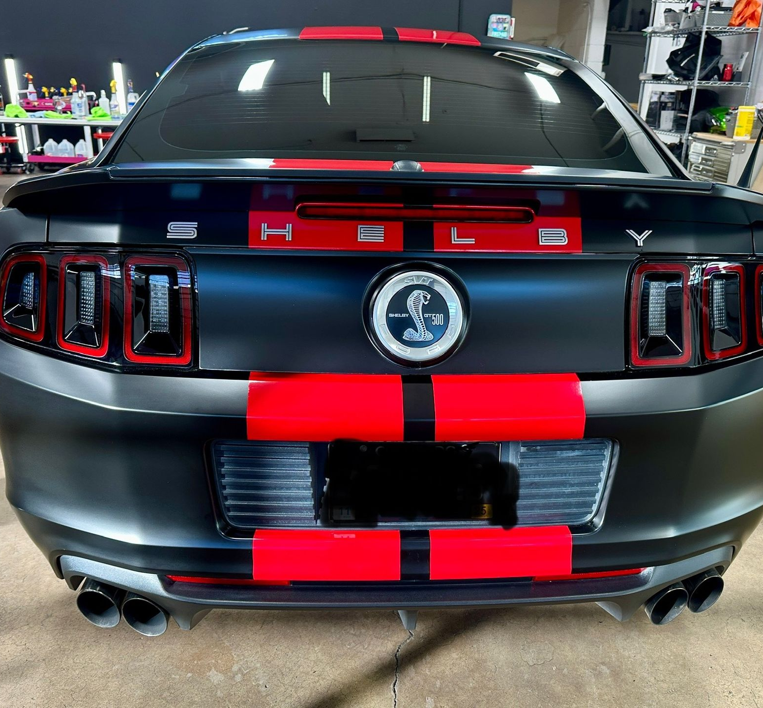 Rear view of a matte black Ford Mustang with red racing stripes in a garage