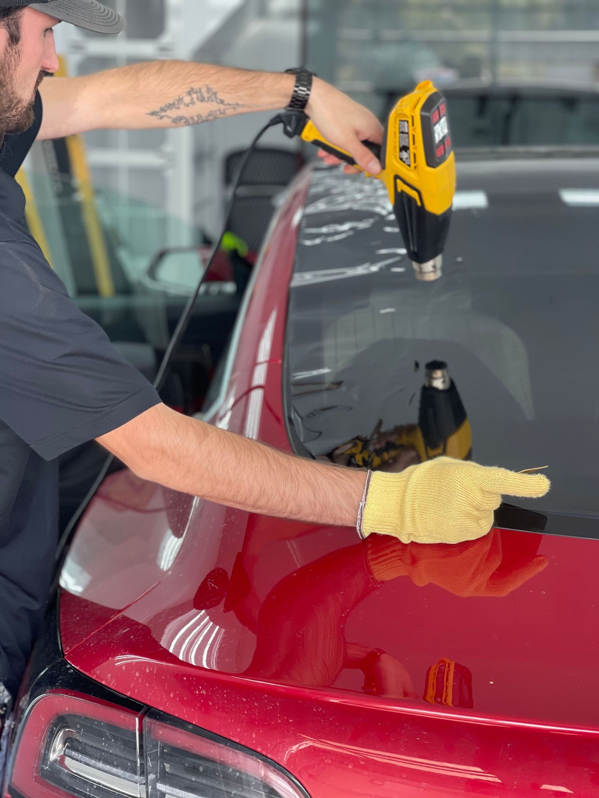A technician uses a heat gun to apply window tint to the rear windshield of a red car.