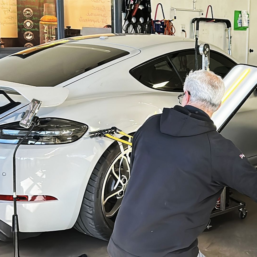 A technician performs paintless dent repair on a car panel using a specialized light board in a garage setting.