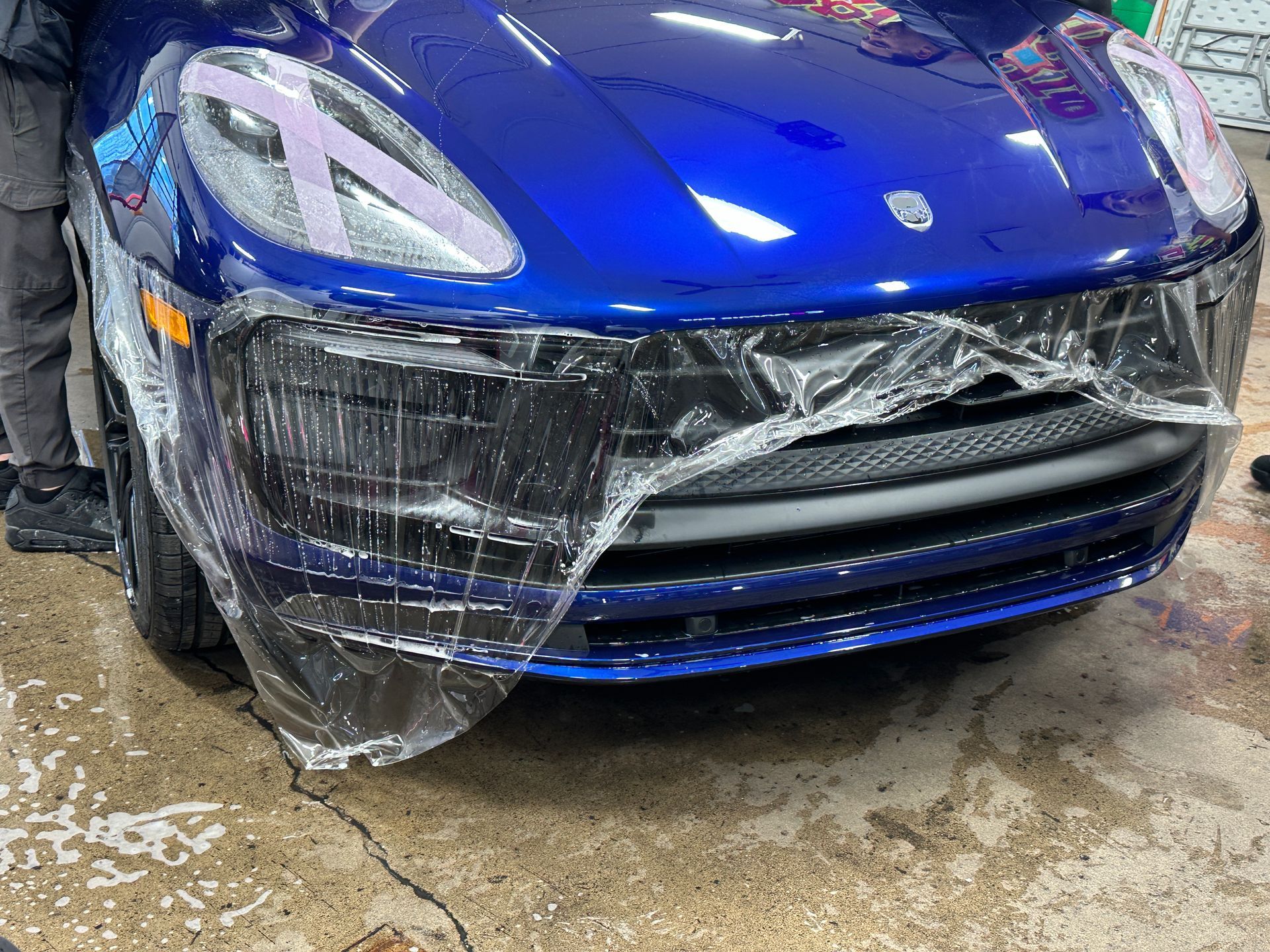 A person uses a yellow squeegee to apply clear paint protection film to the burgundy rear fender of a Porsche Targa.