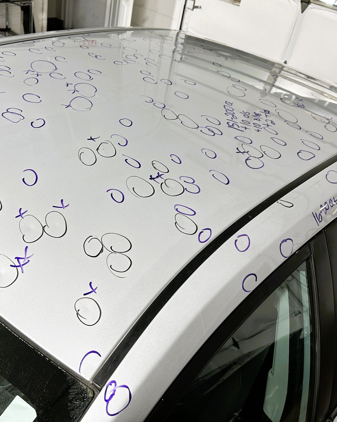 A technician uses a tool to repair small dents on a blue car hood under bright workshop lights.