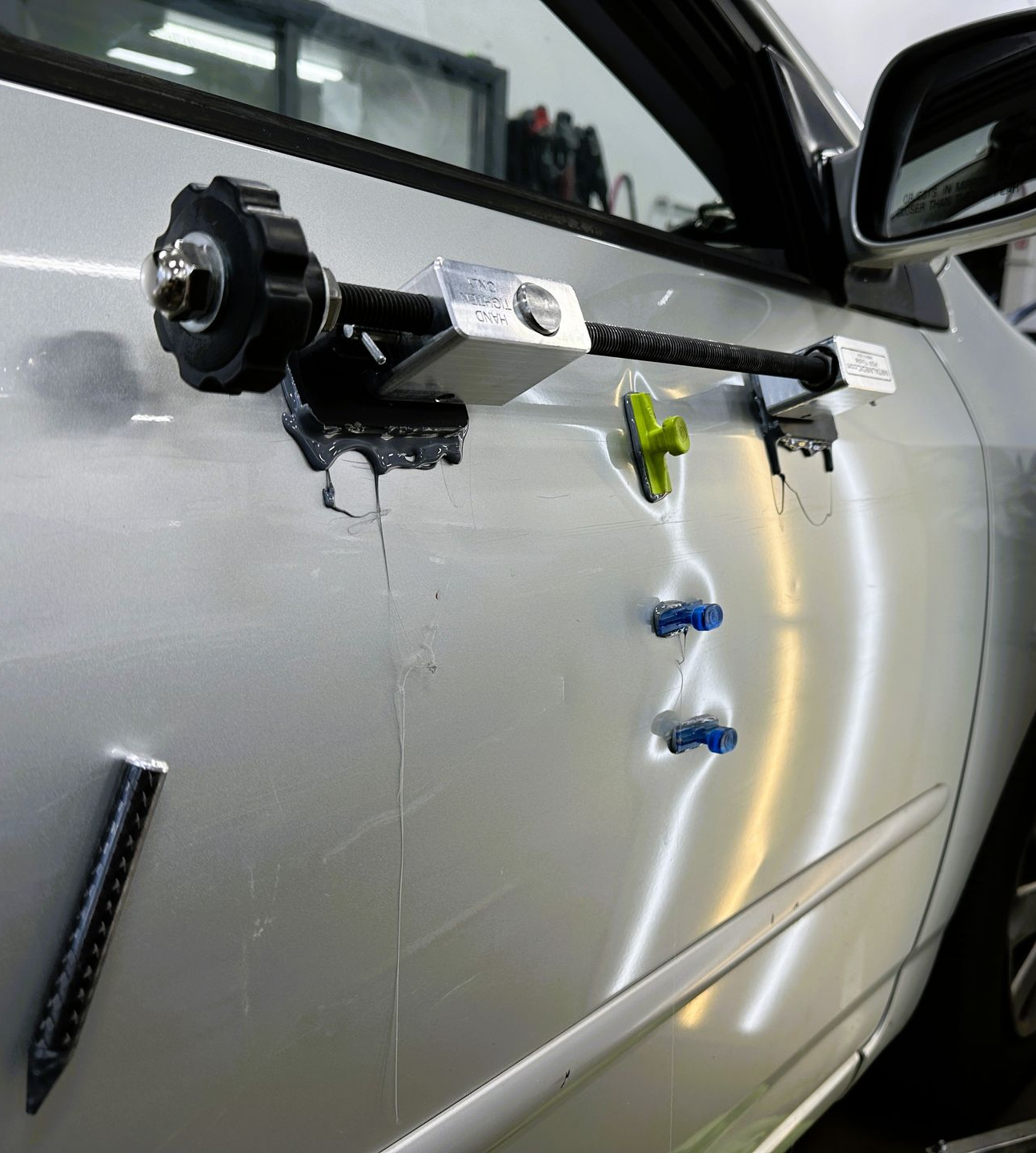 A technician uses a tool to repair a dent on a shiny red car panel, with a yellow reflection board in the background.