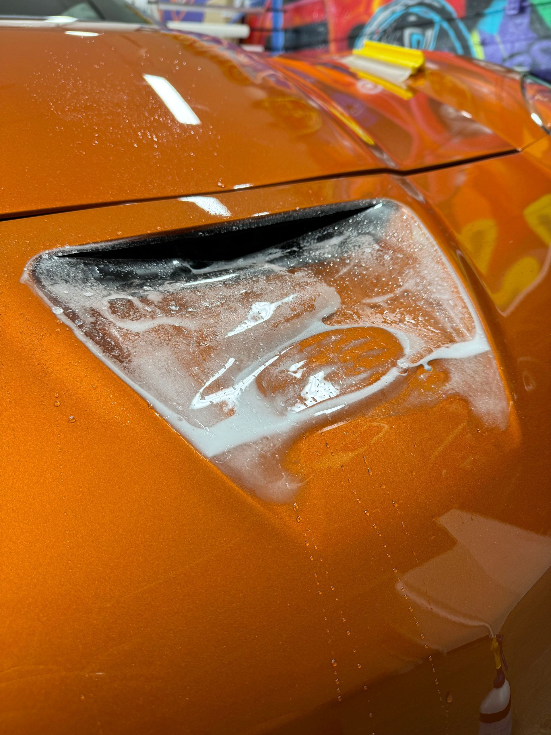 A close-up view of soap suds pooled on the orange hood and air vent of a vehicle during a car wash.