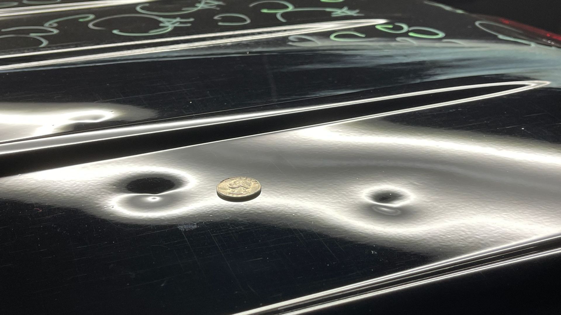 A close-up of a black car hood showing several hail dents, with a coin placed for scale.