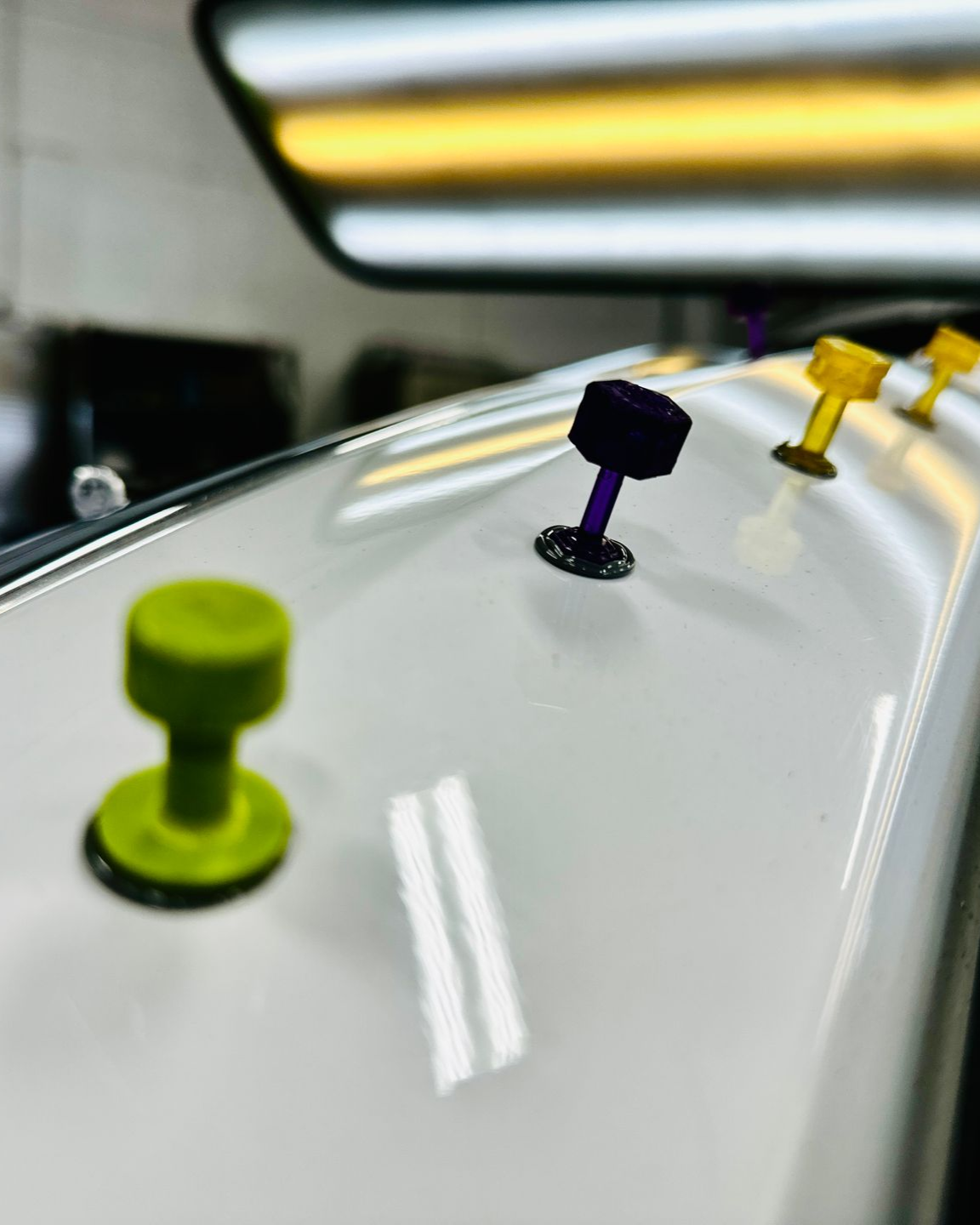 A hand uses a small hammer to perform paintless dent repair on a car hood, illuminated by bright LED workshop lights.