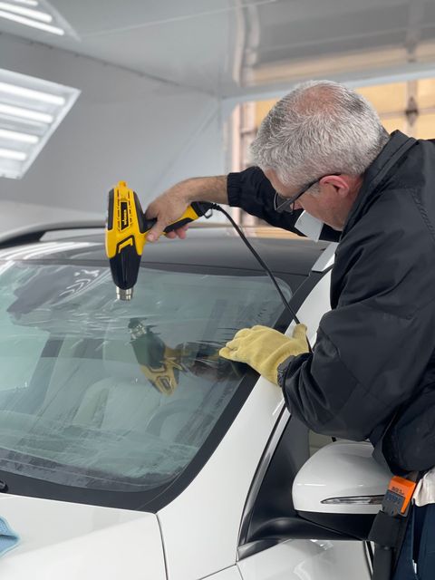 A technician uses a yellow heat gun to apply window tint to the windshield of a white vehicle inside a bright shop.