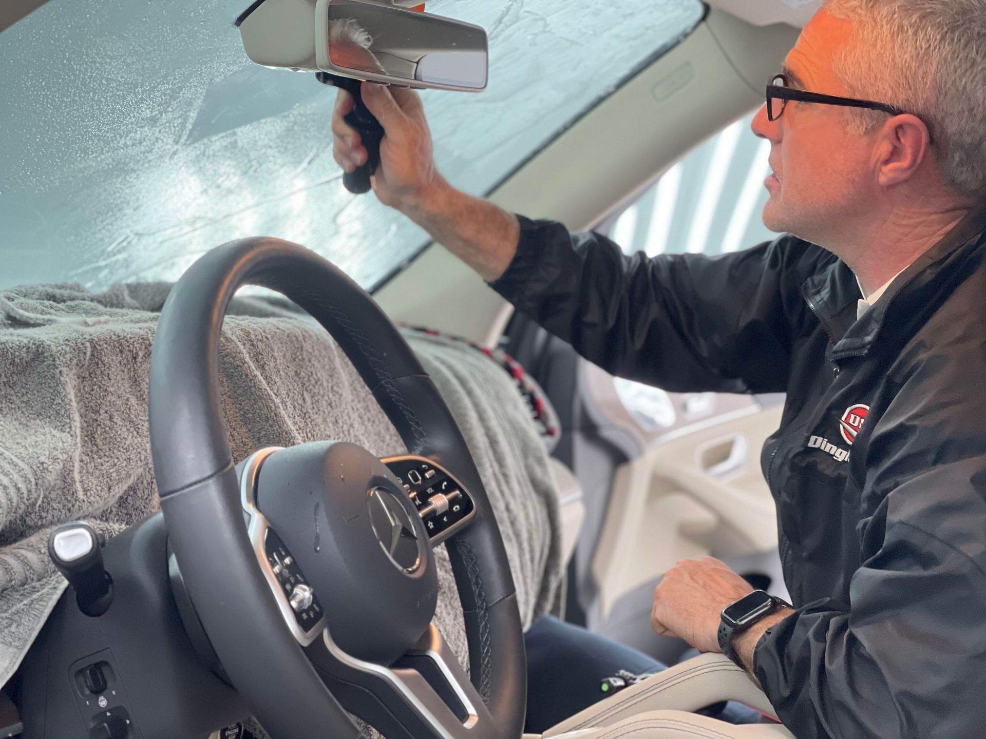 A technician in a black jacket adjusts the rearview mirror inside a car with a Mercedes-Benz steering wheel.