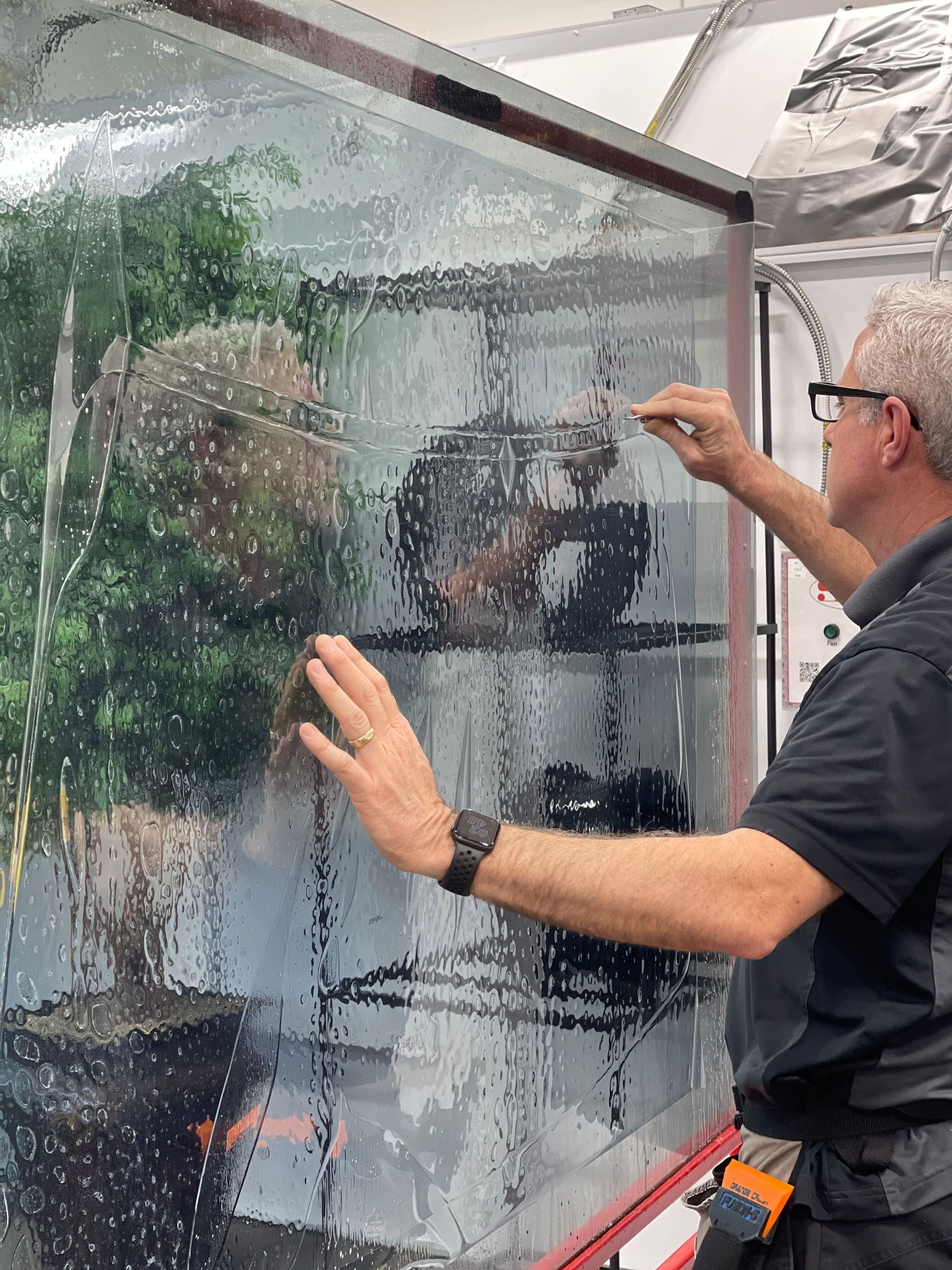 A person wipes a glass pane covered in water droplets with a cleaning tool, revealing a potted plant behind it.