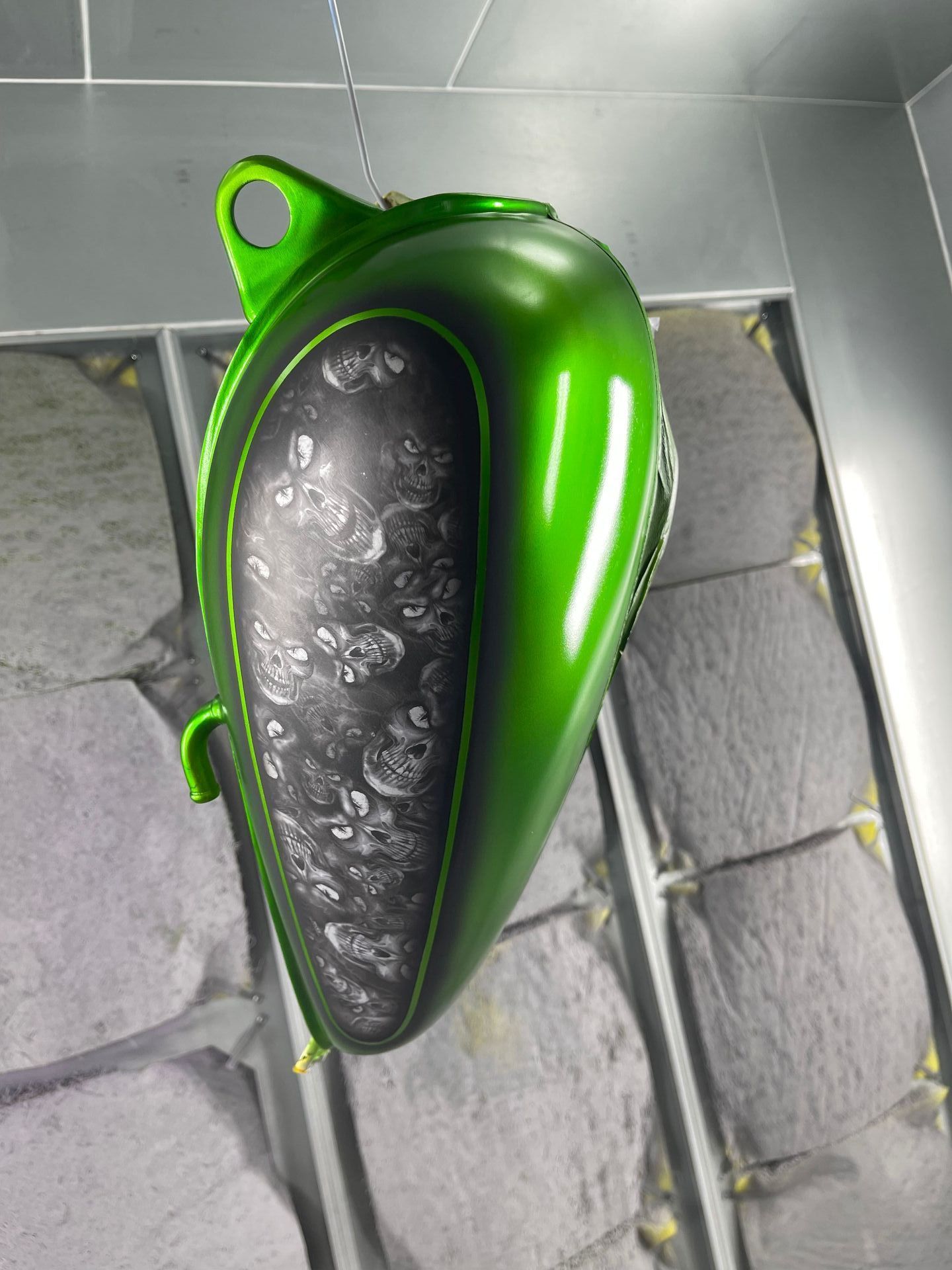 A glossy green motorcycle fuel tank hanging in a paint booth, featuring a skull-patterned inlay on the side panel.