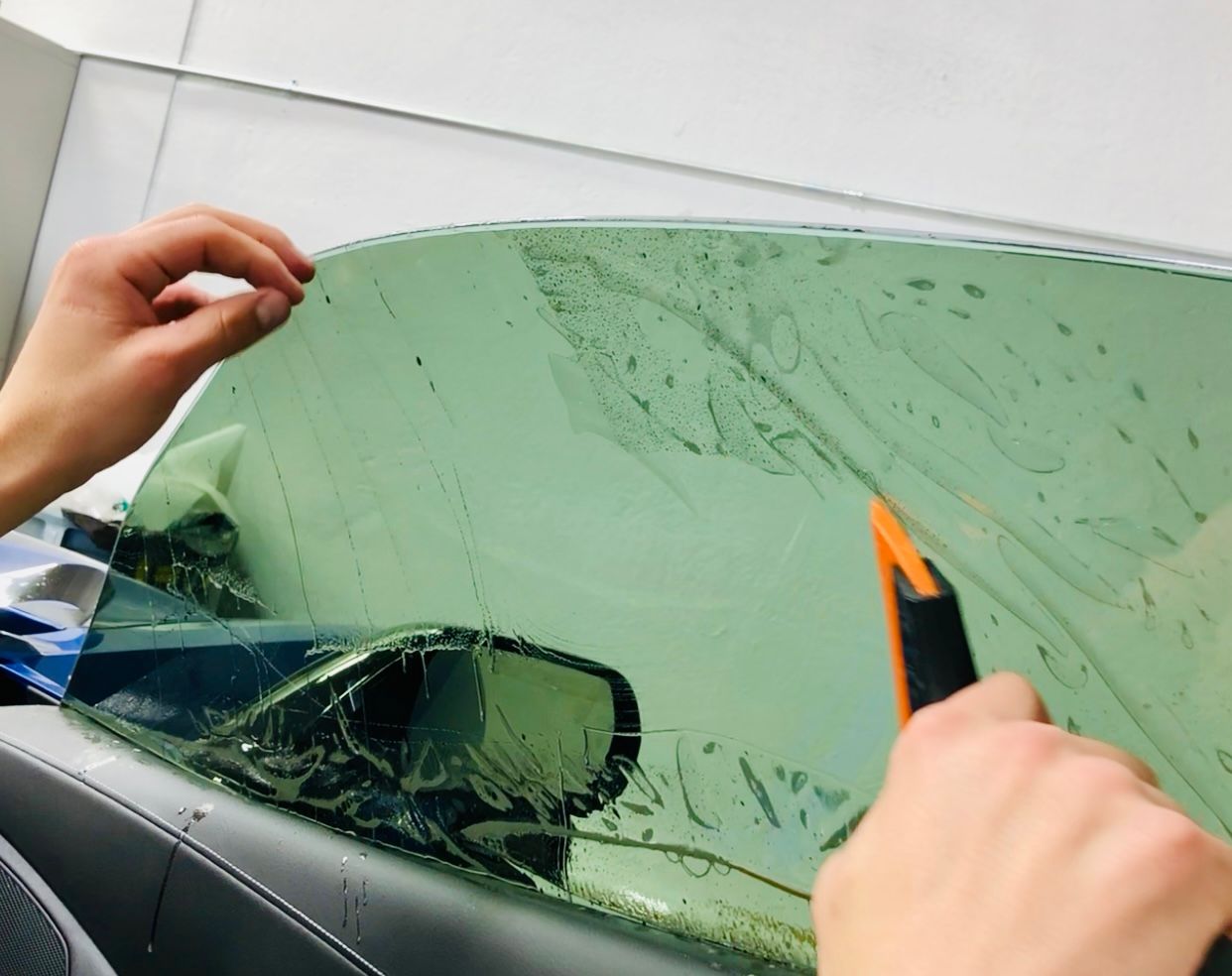 Hands peel green-tinted film from a car windshield with a utility knife.