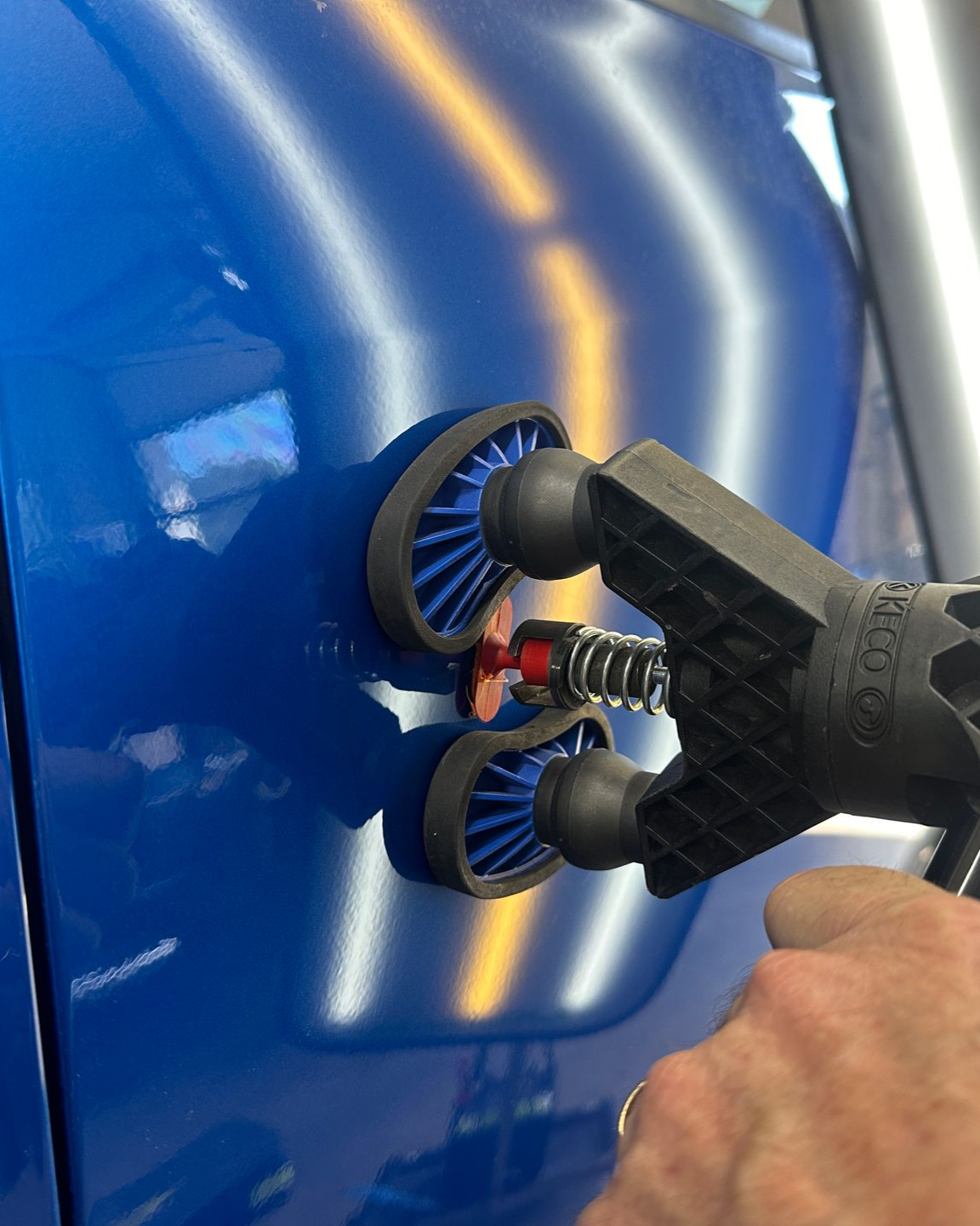 A hand uses a slide hammer tool with a blue glue tab to pull a dent out of a red car panel.