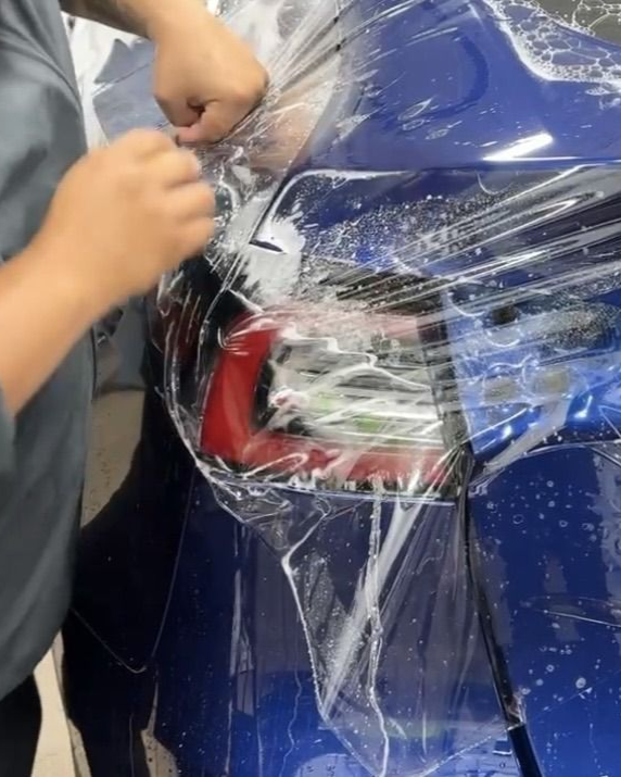 Hands apply a clear protective film to a vehicle headlight using a bright pink squeegee.