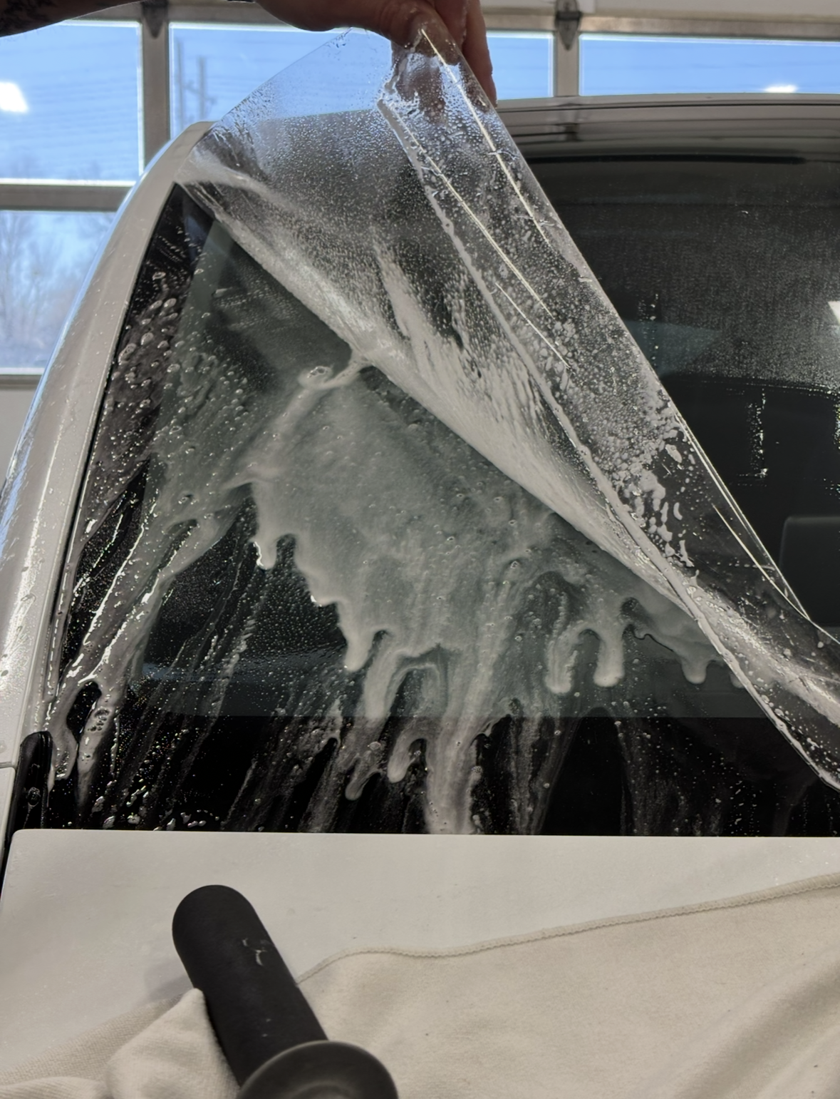 A person applying a clear, soapy window tint film to the rear glass of a vehicle inside a garage.