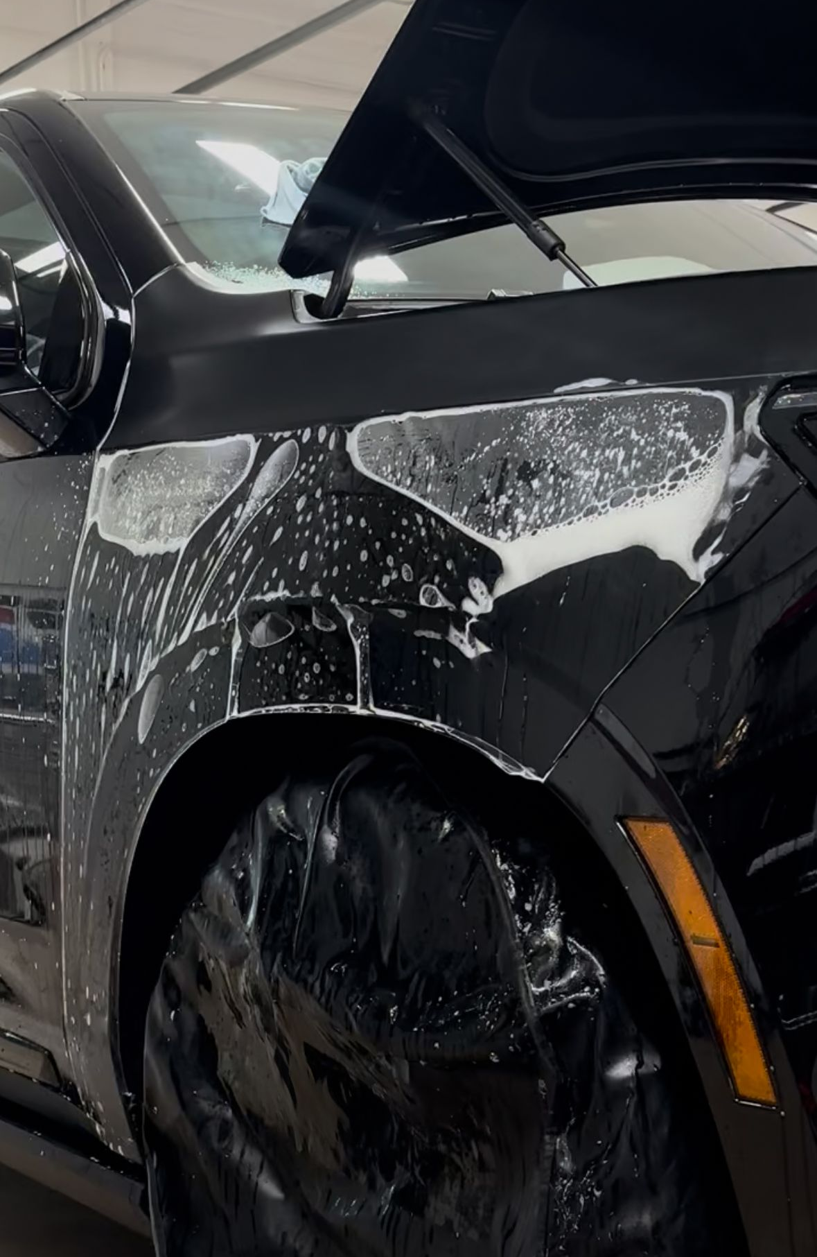A black car fender covered in white cleaning foam during a detailing process, with the wheel masked by plastic wrap.