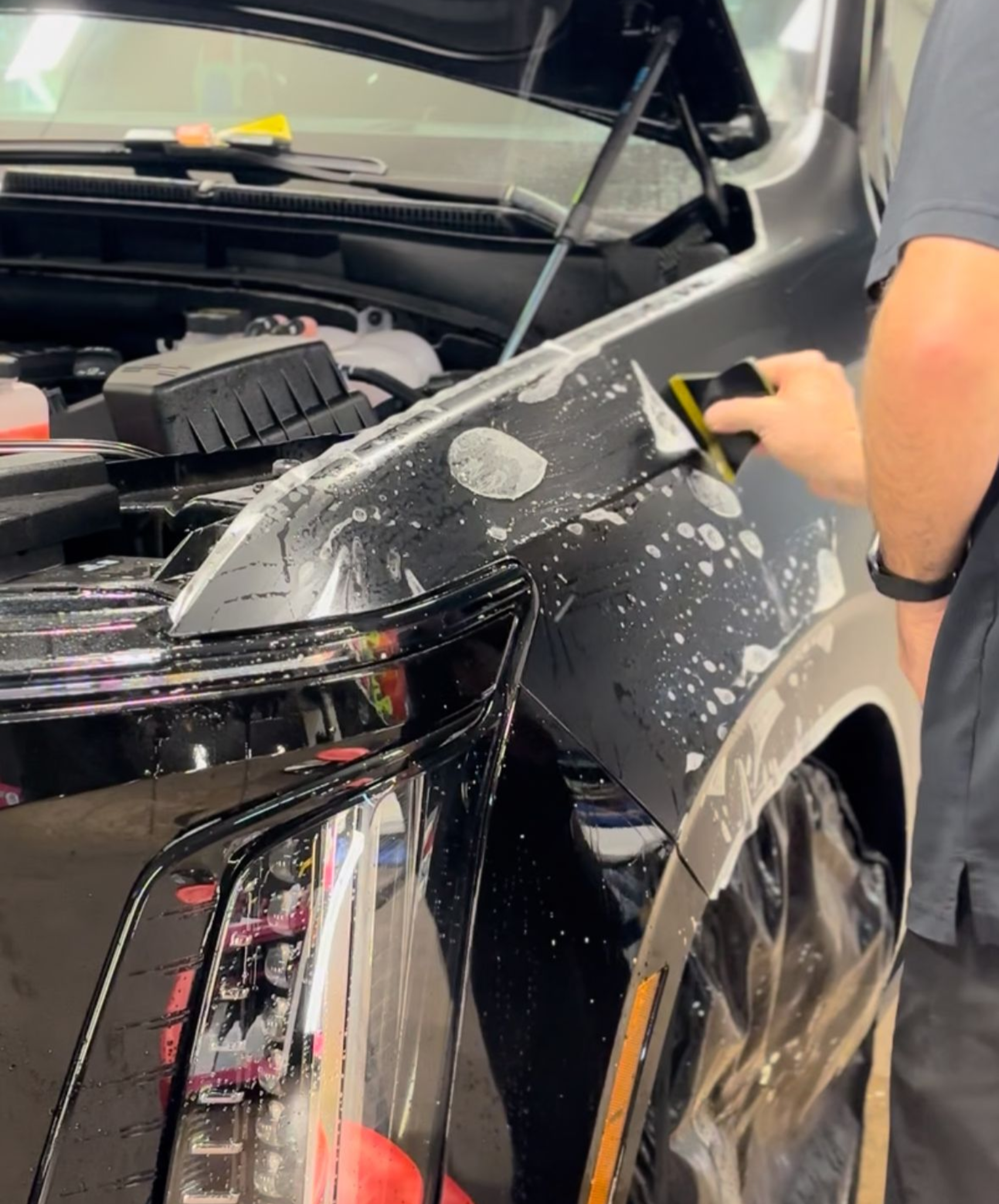 A technician uses a squeegee to apply a clear protective film to the fender of a dark car in a garage.