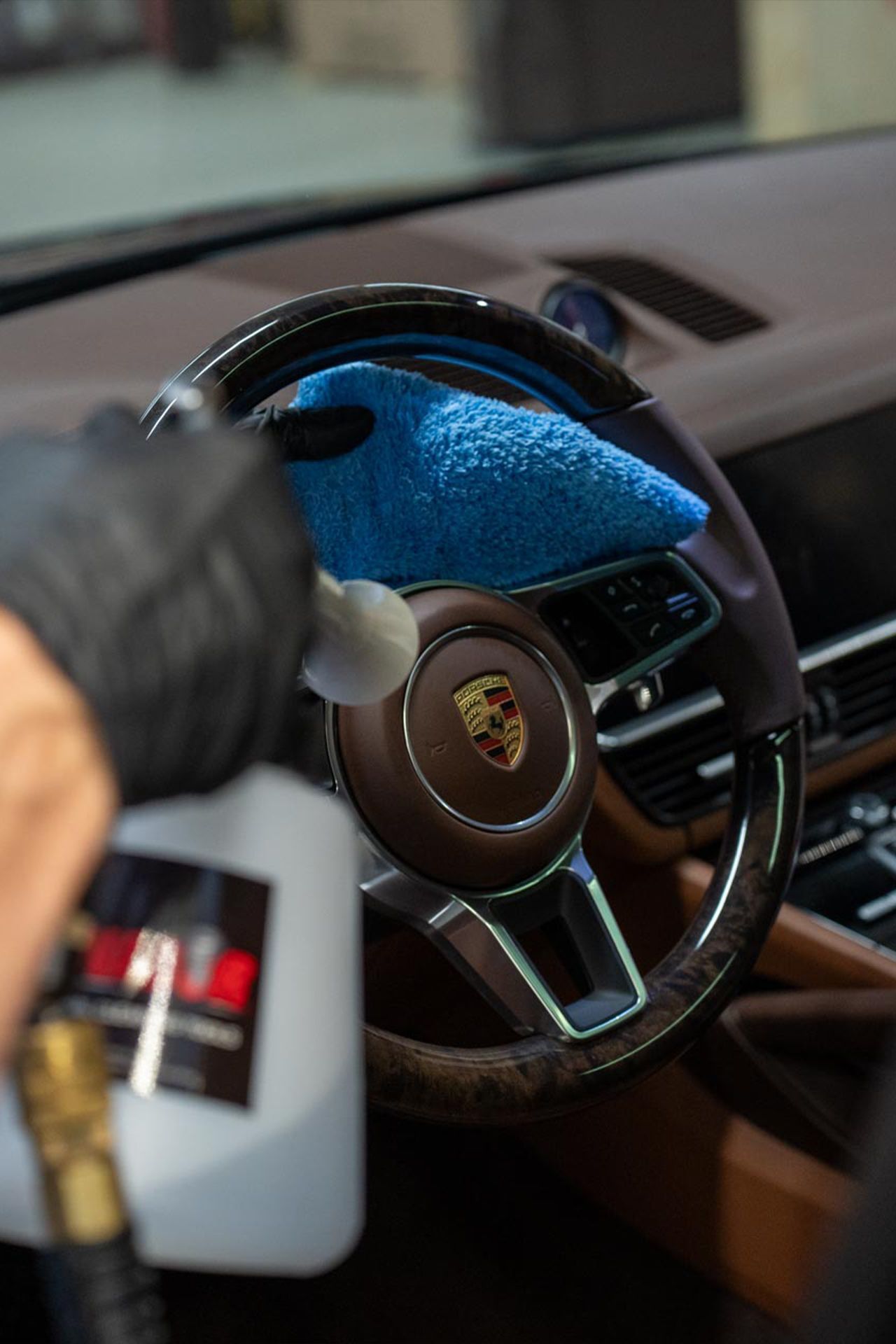 A hand in a black glove holds a spray bottle and blue cloth, cleaning the steering wheel of a brown leather Porsche interior.