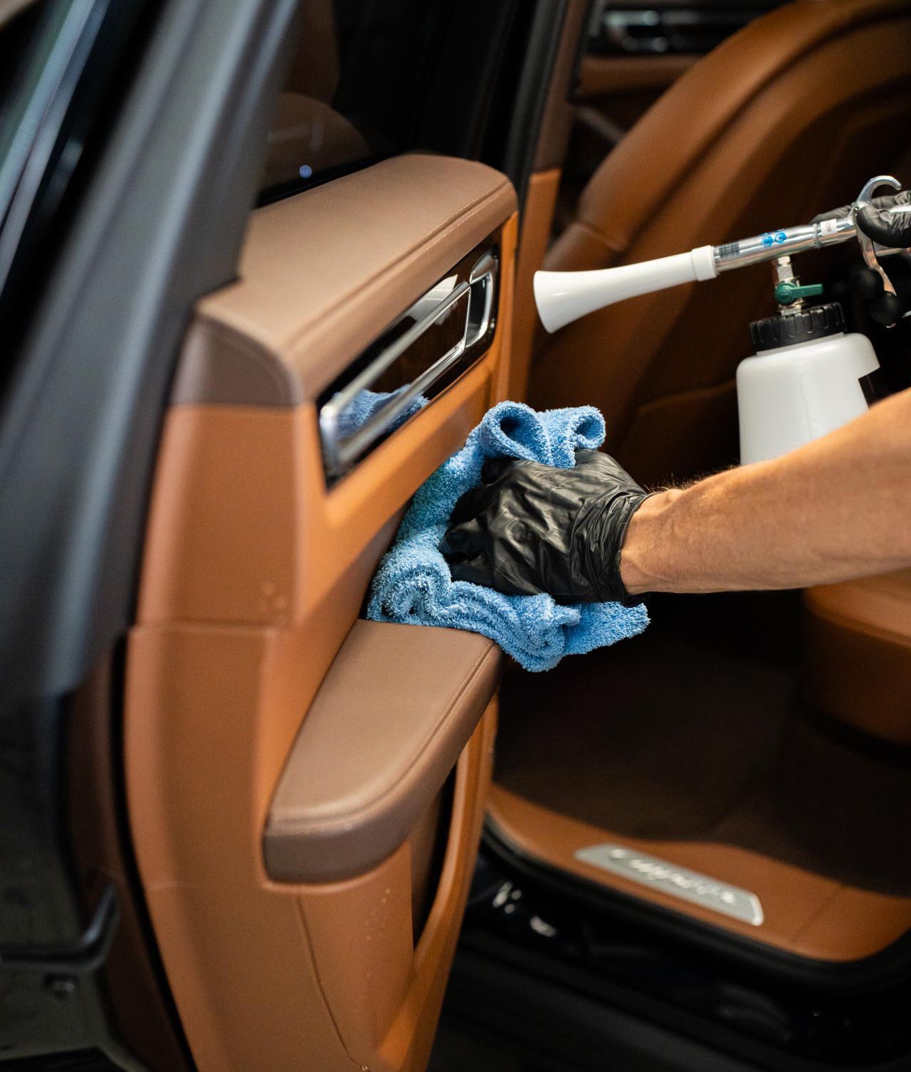 A person wearing a black glove uses a blue microfiber cloth and a spray tool to clean a brown leather car door panel.