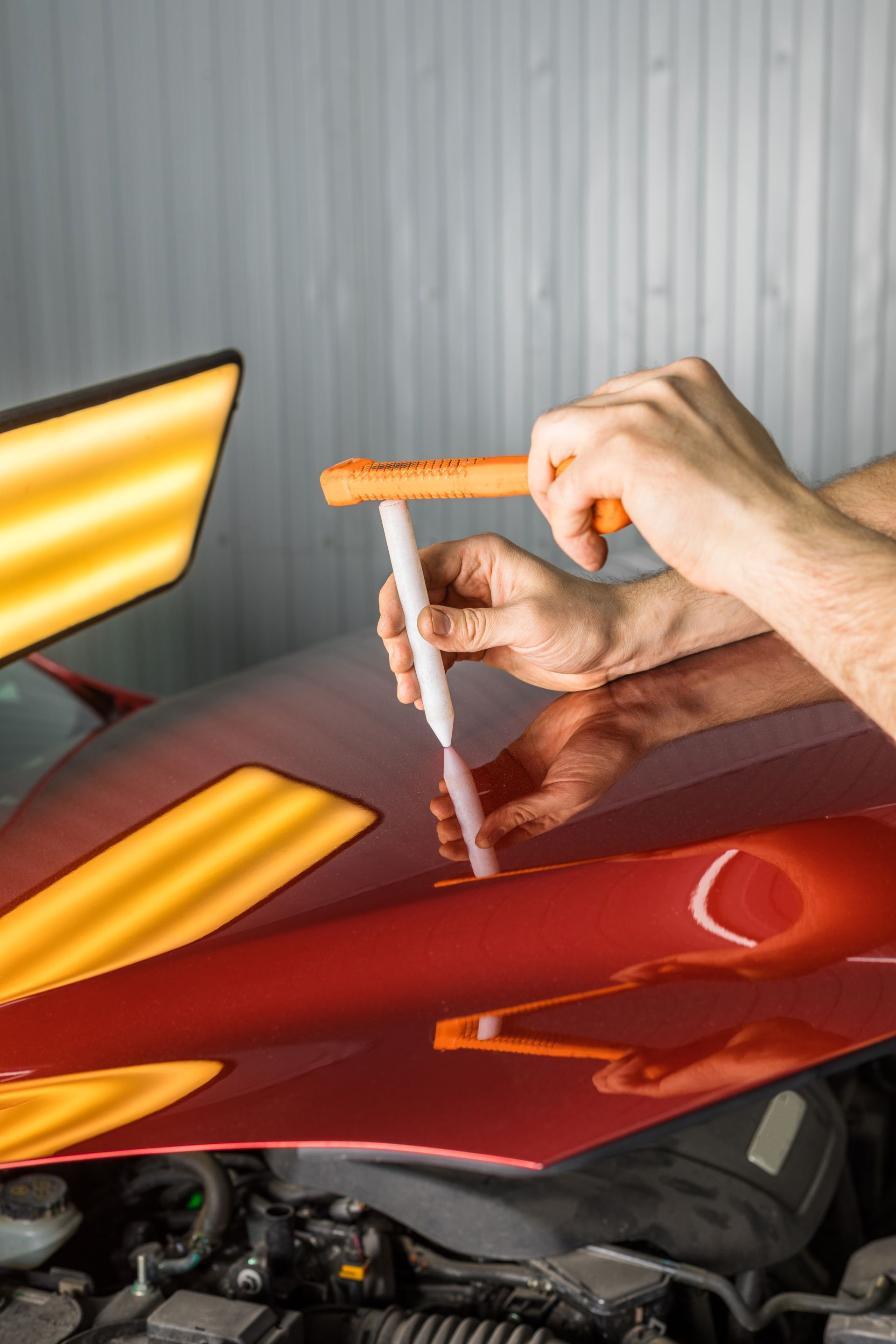 A technician uses a tool to perform paintless dent repair on the red hood of a vehicle under a professional light panel.