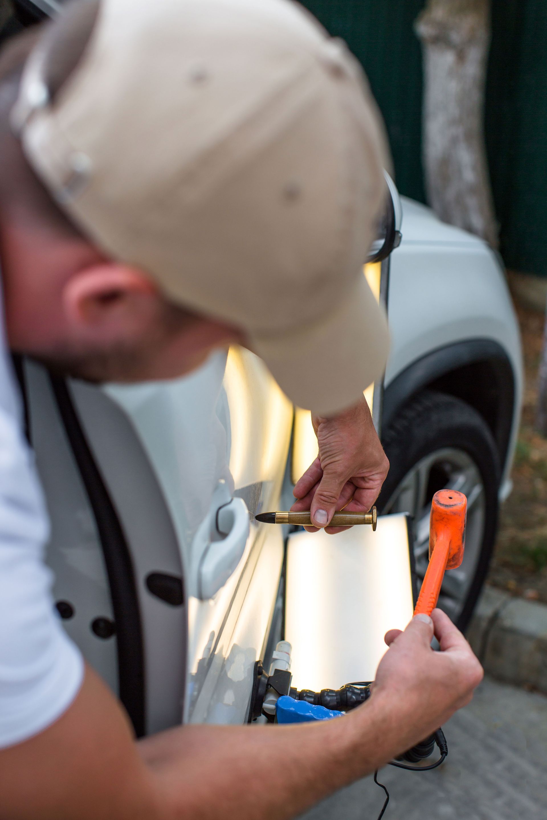 A person uses a punch tool and mallet to perform paintless dent repair on a white car door with a light board.